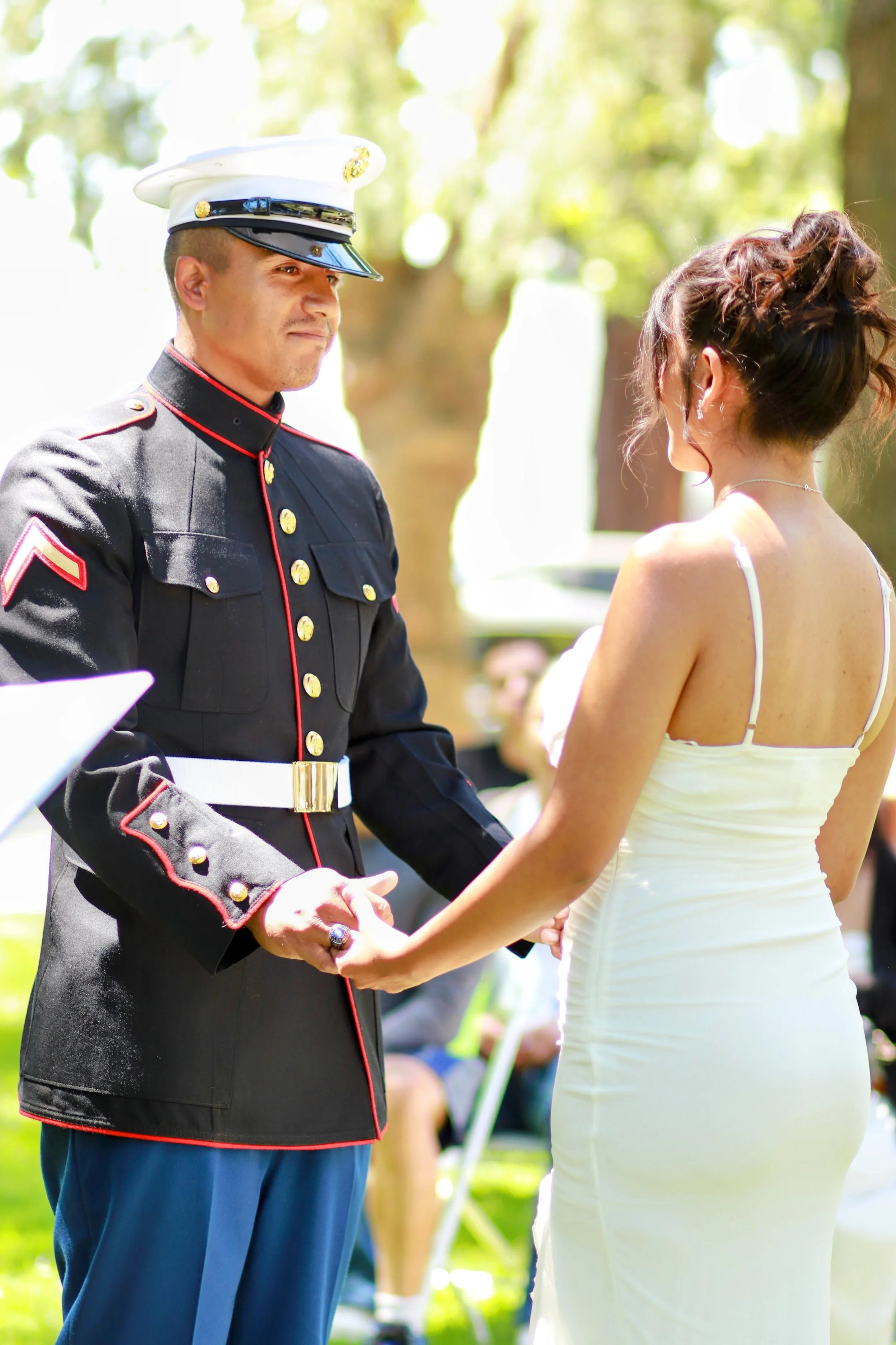 A man in a U.S. Marine dress uniform holding hands with a woman in a white wedding dress, outdoors in a bright, sunny park setting.