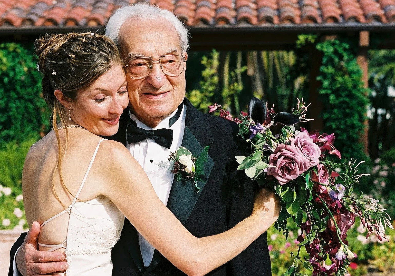 A young woman in a white dress hugging an elderly grandparent in a tuxedo, holding a large bouquet of flowers, outdoors with greenery and a building with a tiled spanish tile roof in the background.