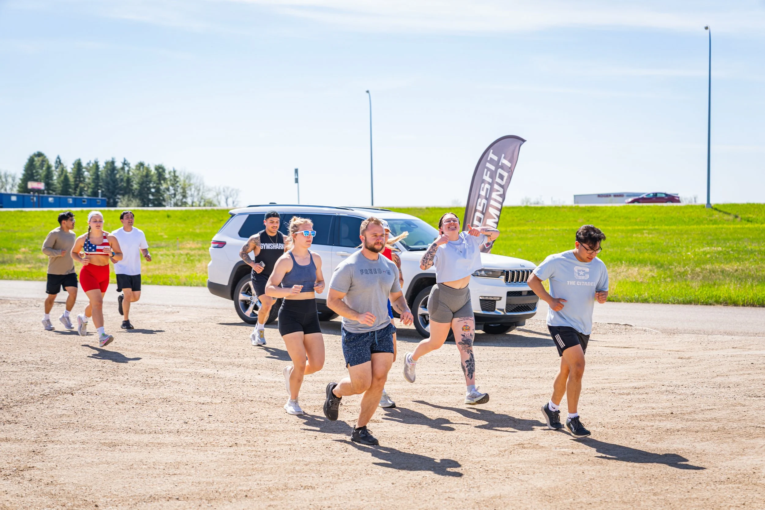 Group of people running outdoors on a gravel path during daytime, with a grassy area, parked cars, and a flag that reads 'Fitness Mind' in the background.