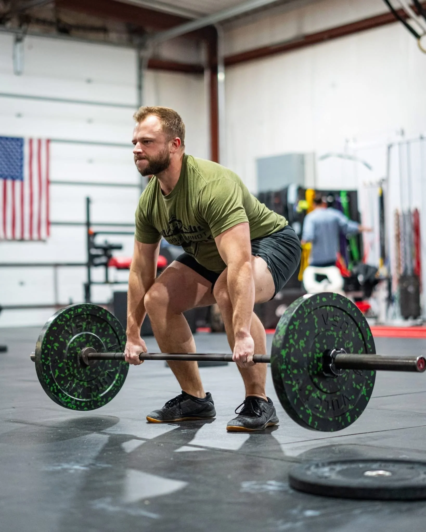 A man wearing a green shirt and black shorts lifting a barbell with green and black weight plates inside a gym.