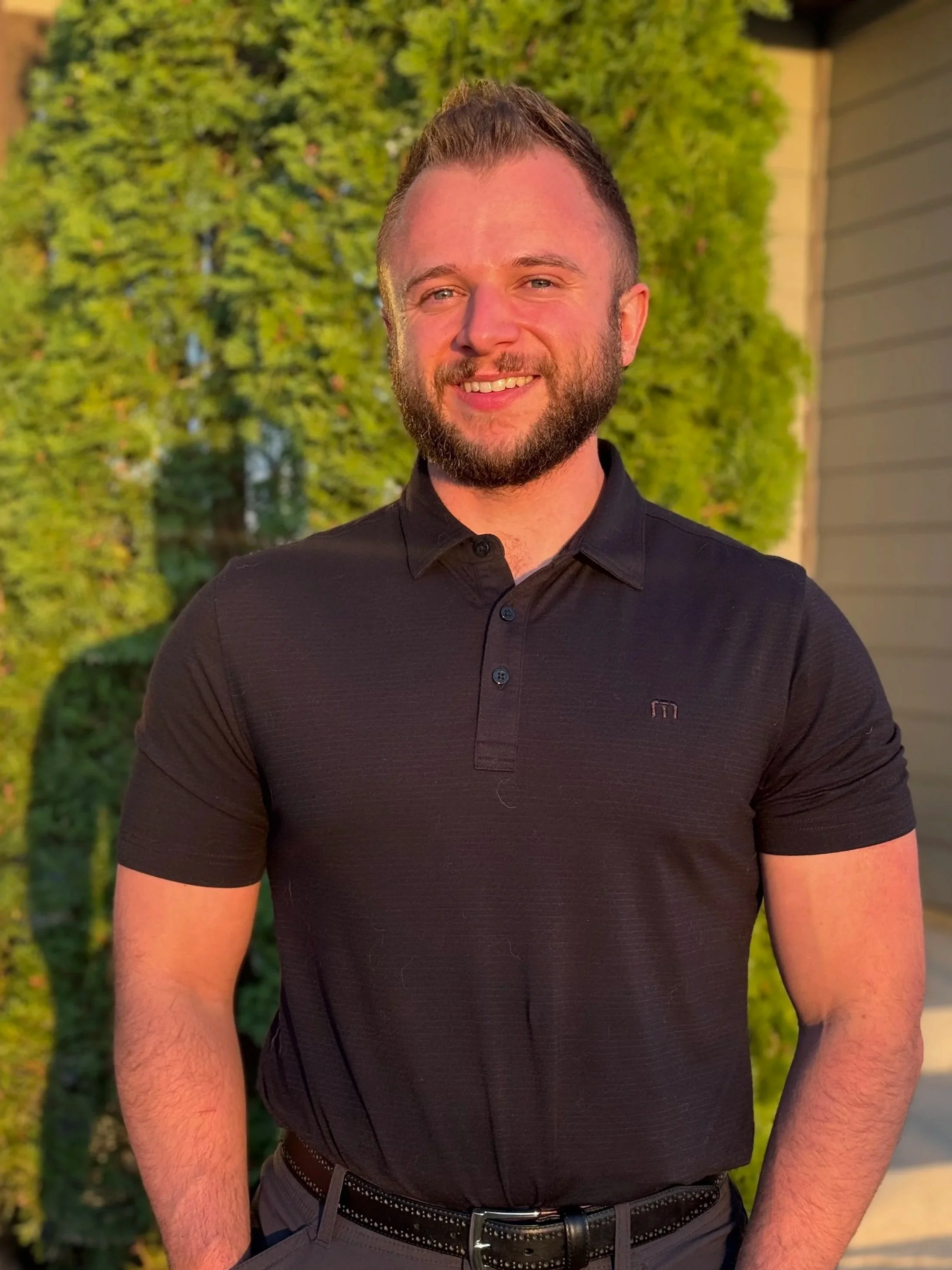 A man with a beard and short hairstyle smiling outdoors during sunset, wearing a black polo shirt and gray pants, with greenery and a house in the background.