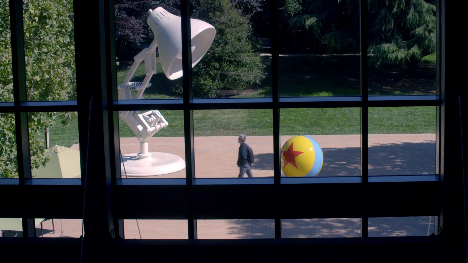 View through window of a large white desk lamp with a spring, a yellow and blue beach ball with a red star, and a person walking outdoors near a brick pathway and green trees.