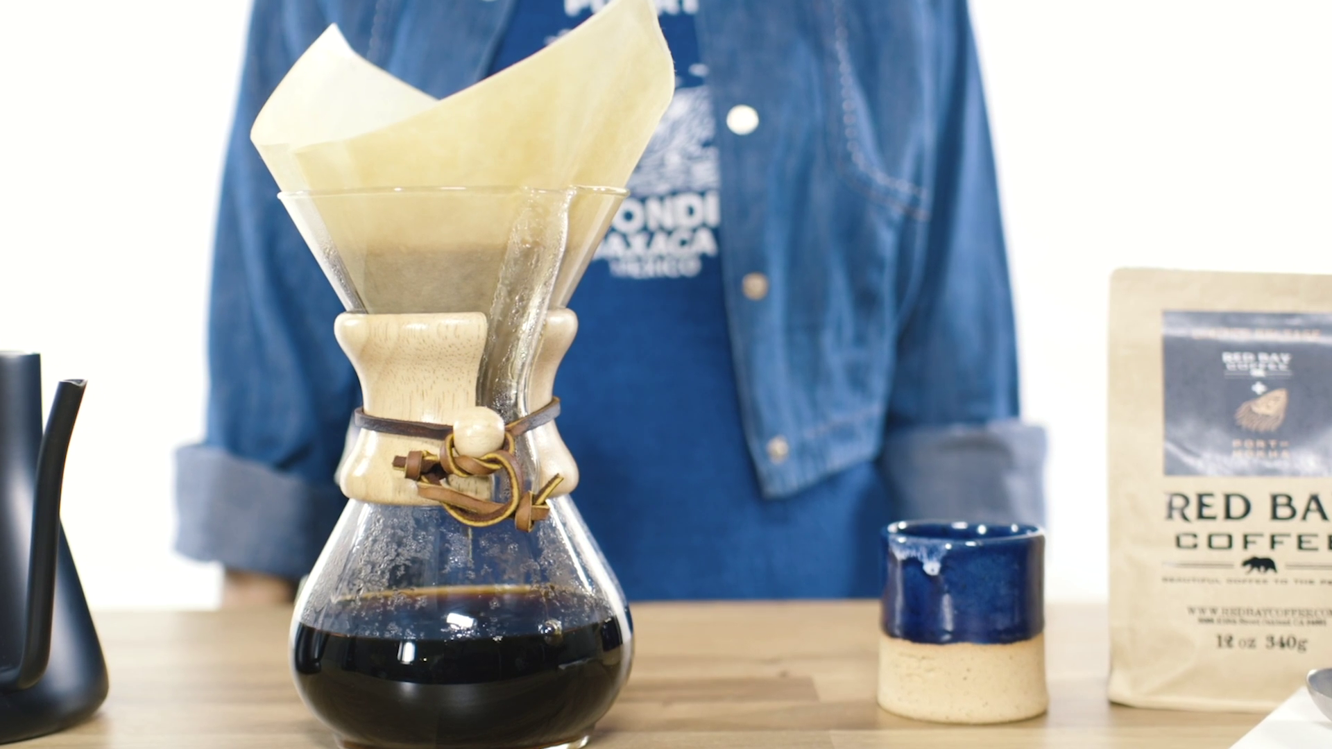 Pour-over coffee brewing setup with a glass dripper, paper filter, and coffee carafe, on a wooden table, with a person in a blue denim shirt in the background.
