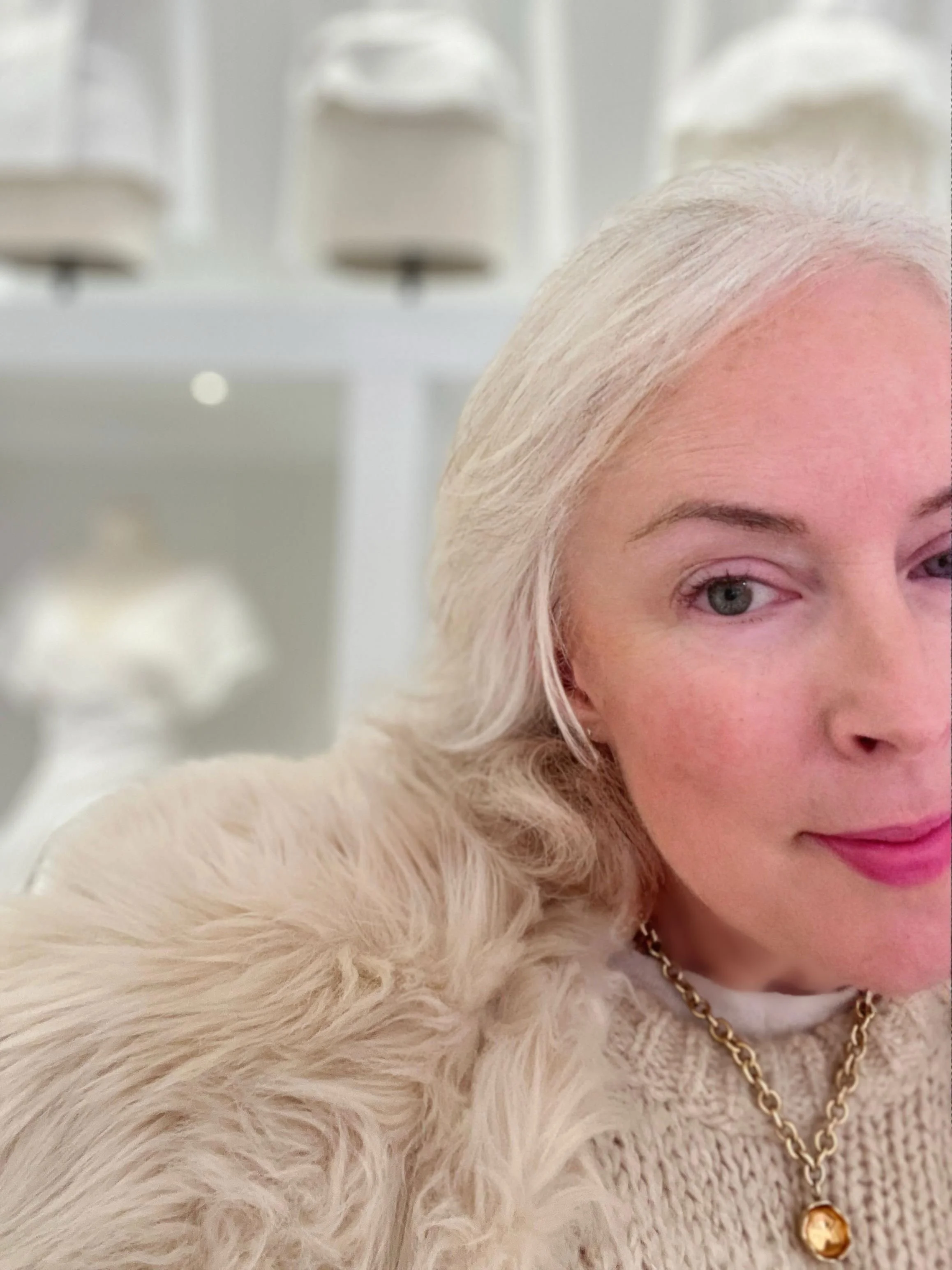 Close-up of Millicent with blonde hair, wearing a gold chain necklace with a large pendant, a white furry jacket, and pink lipstick, at La Galerie Dior featuring white dress forms of the history of Dior desings.