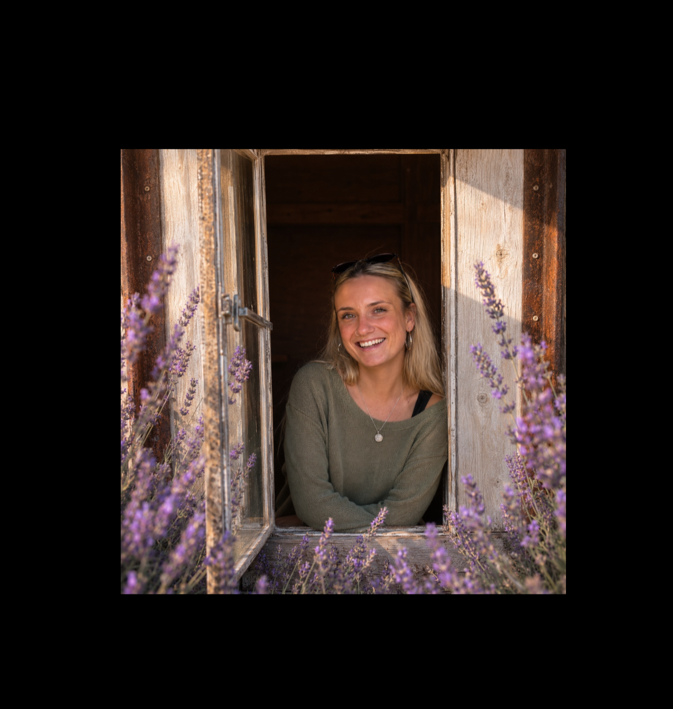 A woman with blonde hair and wearing a green sweater and sunglasses on her head leaning out of a rustic wooden window, smiling, with purple flowers in the foreground.