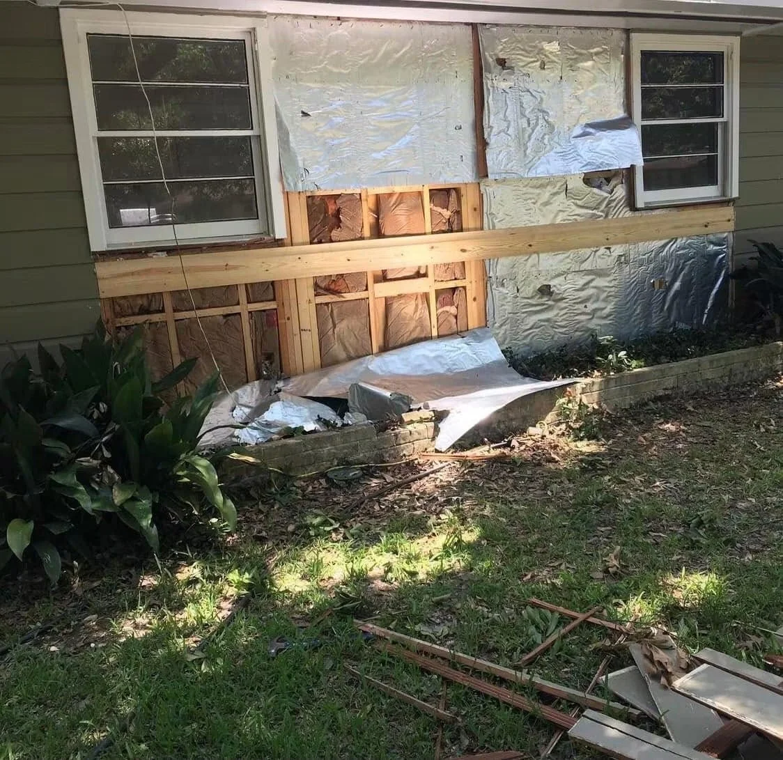 Partially demolished house exterior with insulation and drywall torn down, exposing wooden framing, two windows, and debris on the ground.
