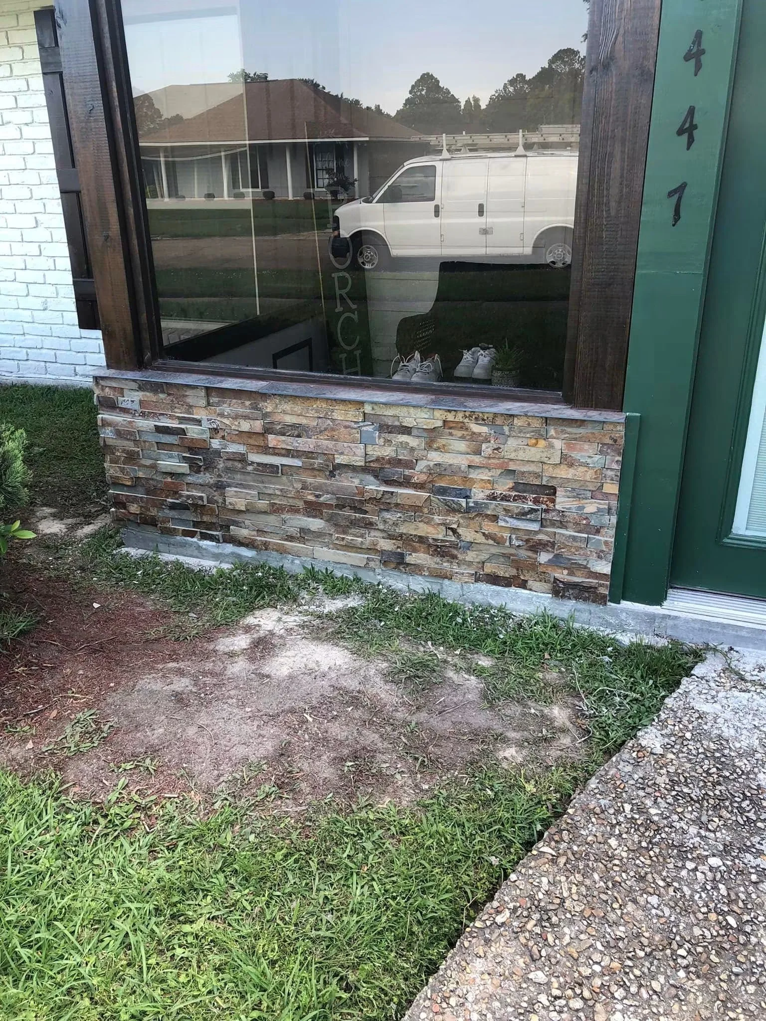 View of a house with a large window, a white van parked outside, a wooden and stone exterior, and a gravel pathway surrounded by grass and plants.