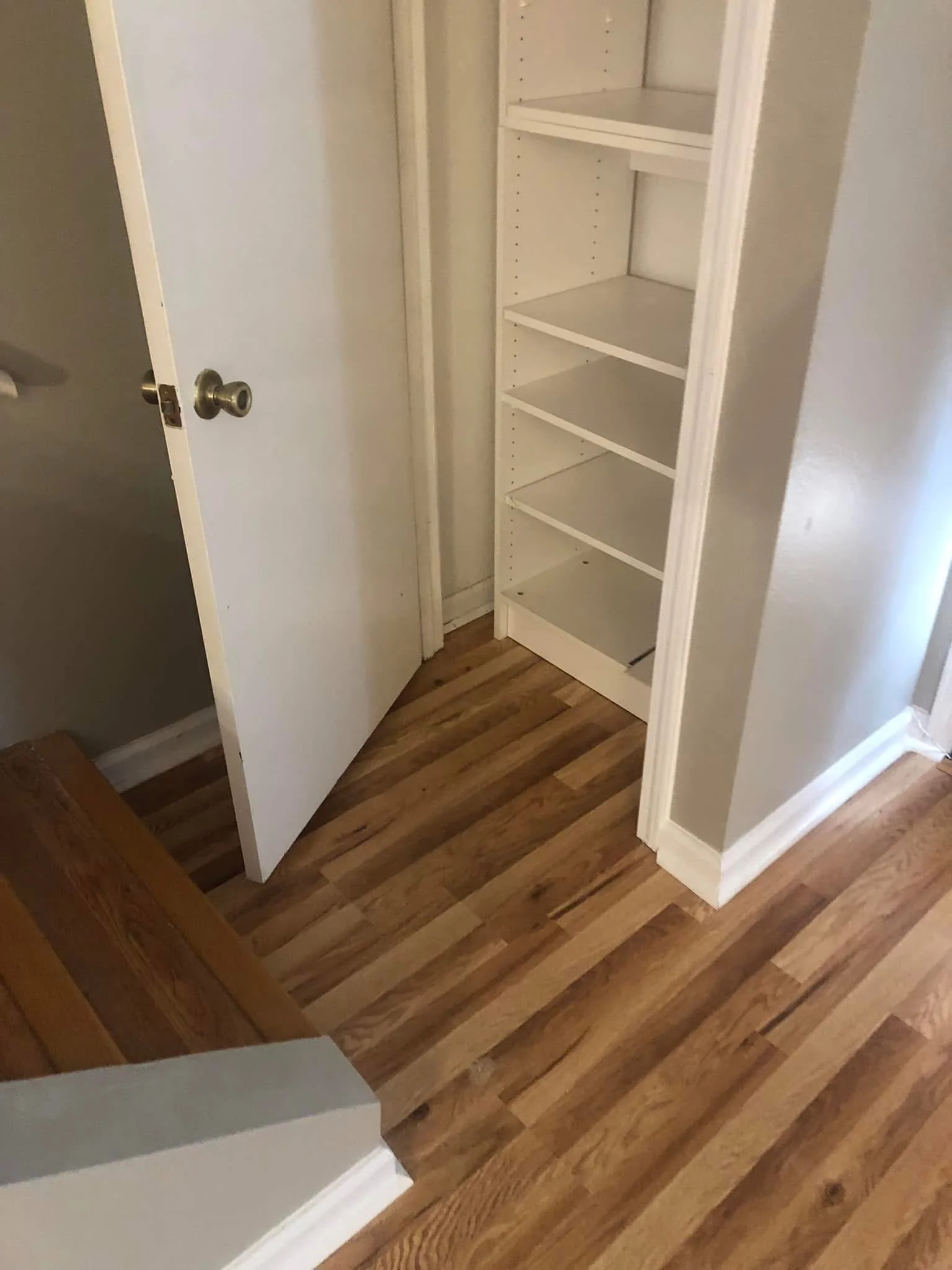 Empty white wooden closet with shelves, open door, built into wall, next to hardwood floor.