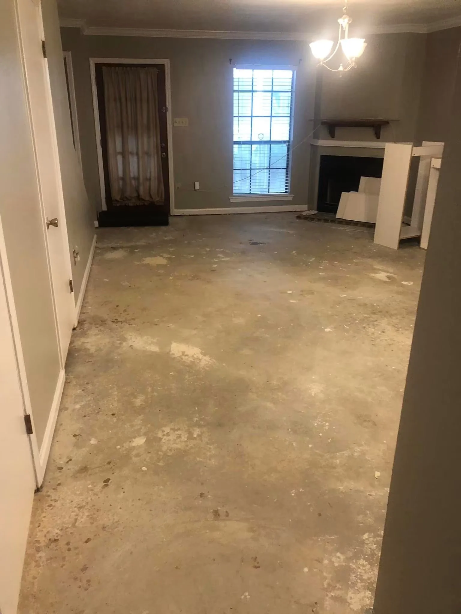 Empty living room with a grey wall, window with blinds, fireplace with a small wooden shelf above, chandelier light fixture, and unfinished floor with patches of debris.