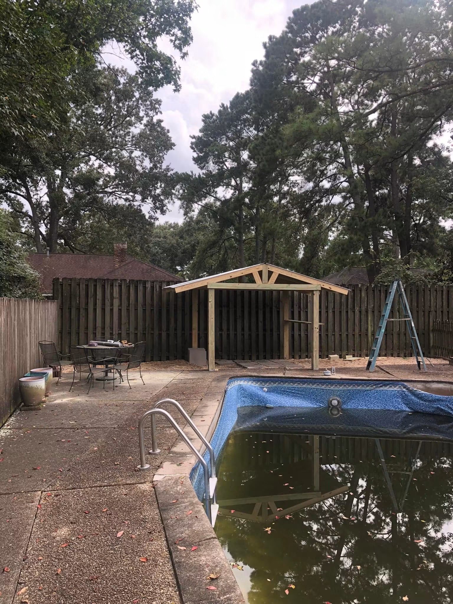 Backyard scene with a fenced area, a small unfinished wooden structure, a ladder, an empty table setting, and a swimming pool with green algae, surrounded by trees and neighboring houses under an overcast sky.