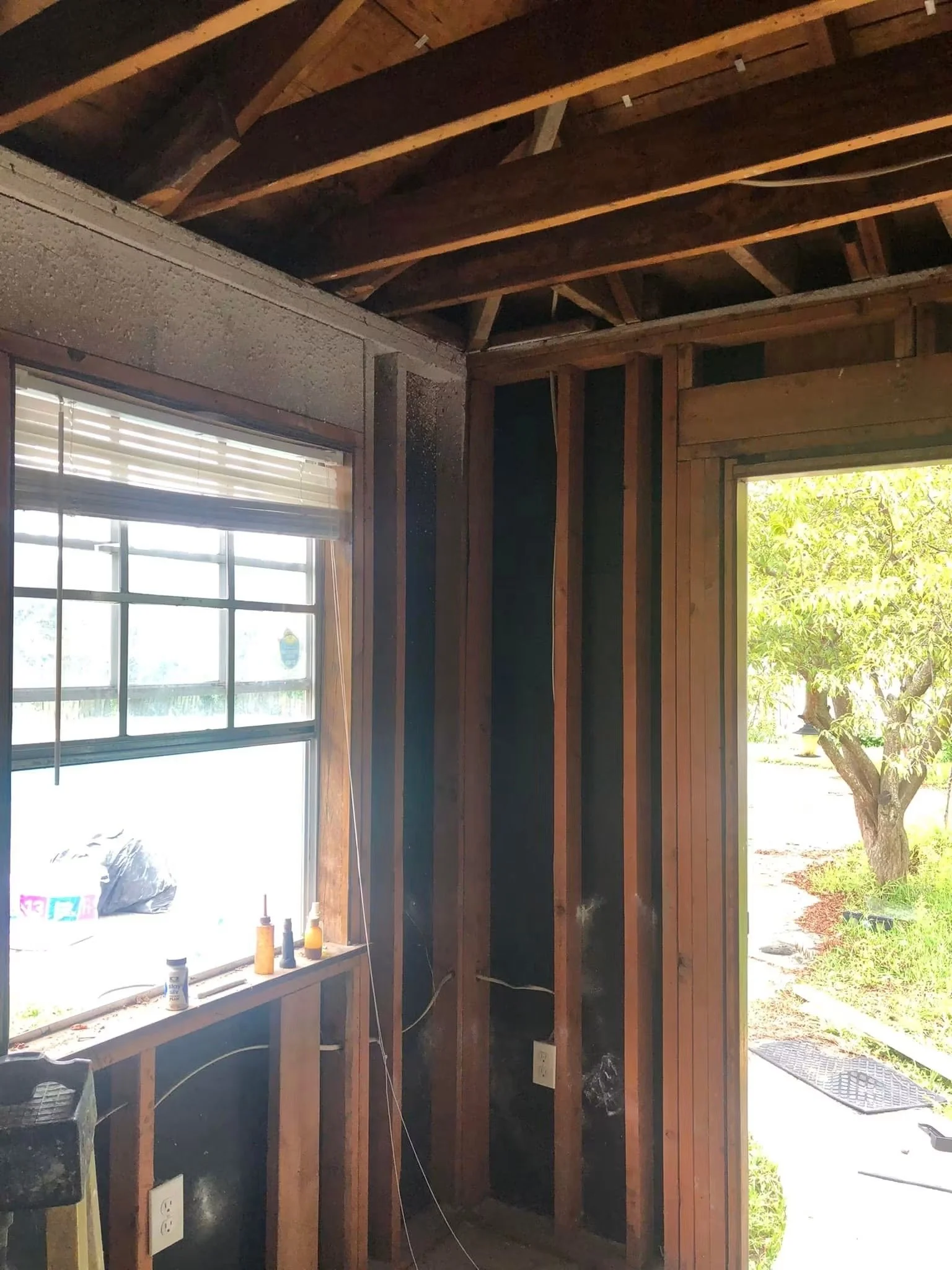 Partially constructed interior of a building with exposed wooden framing and two windows, with construction supplies on the window sill and an outdoor view of a tree and greenery.