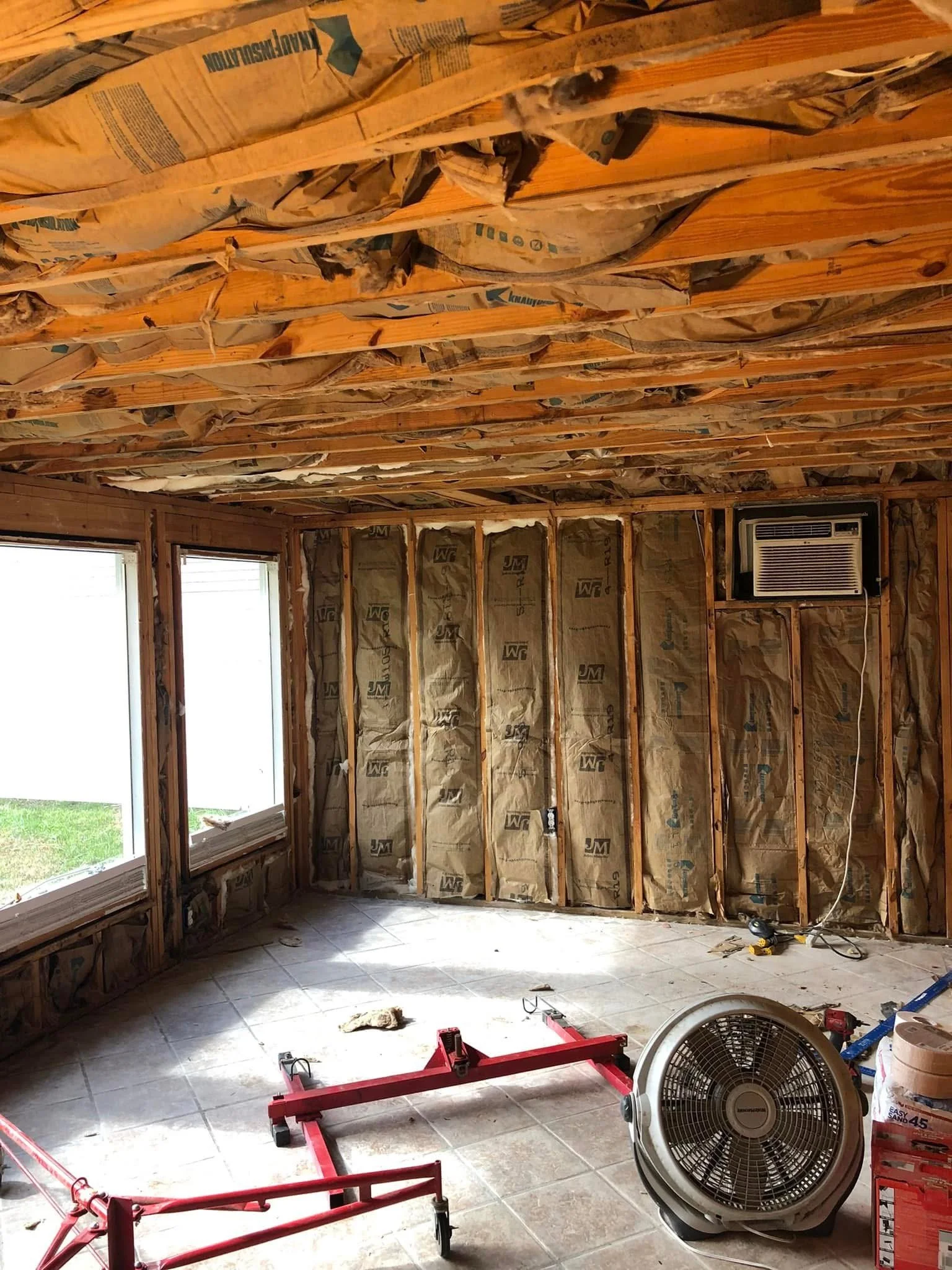 Interior room under renovation with exposed wall studs, insulation, and a mounted window air conditioner. Tools, a fan, and construction materials are scattered across the tiled floor.