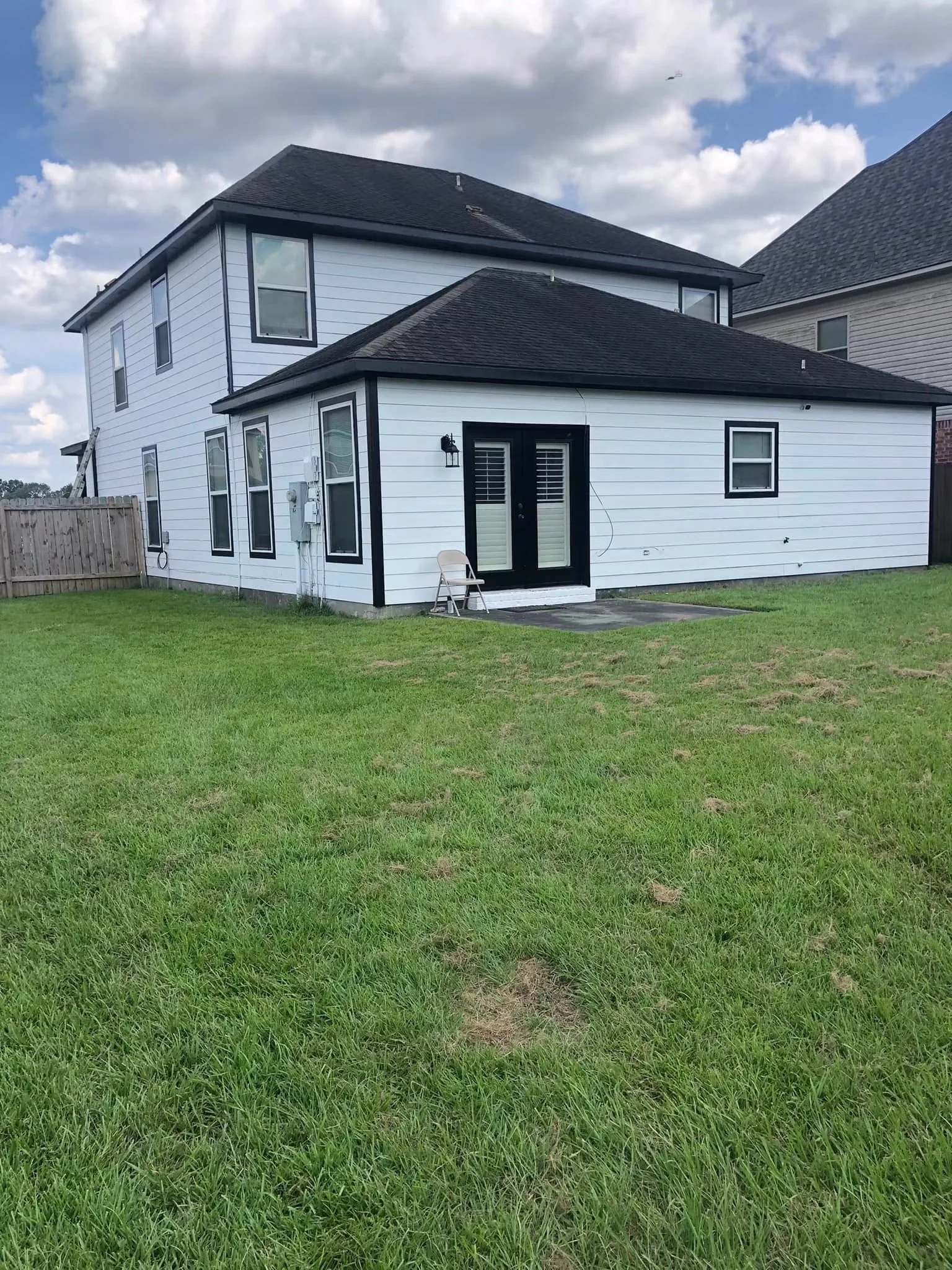 Backyard view of a white house with multiple windows and black trim, a black door, a small patio with a chair, and a fenced yard with green grass under a partly cloudy sky.