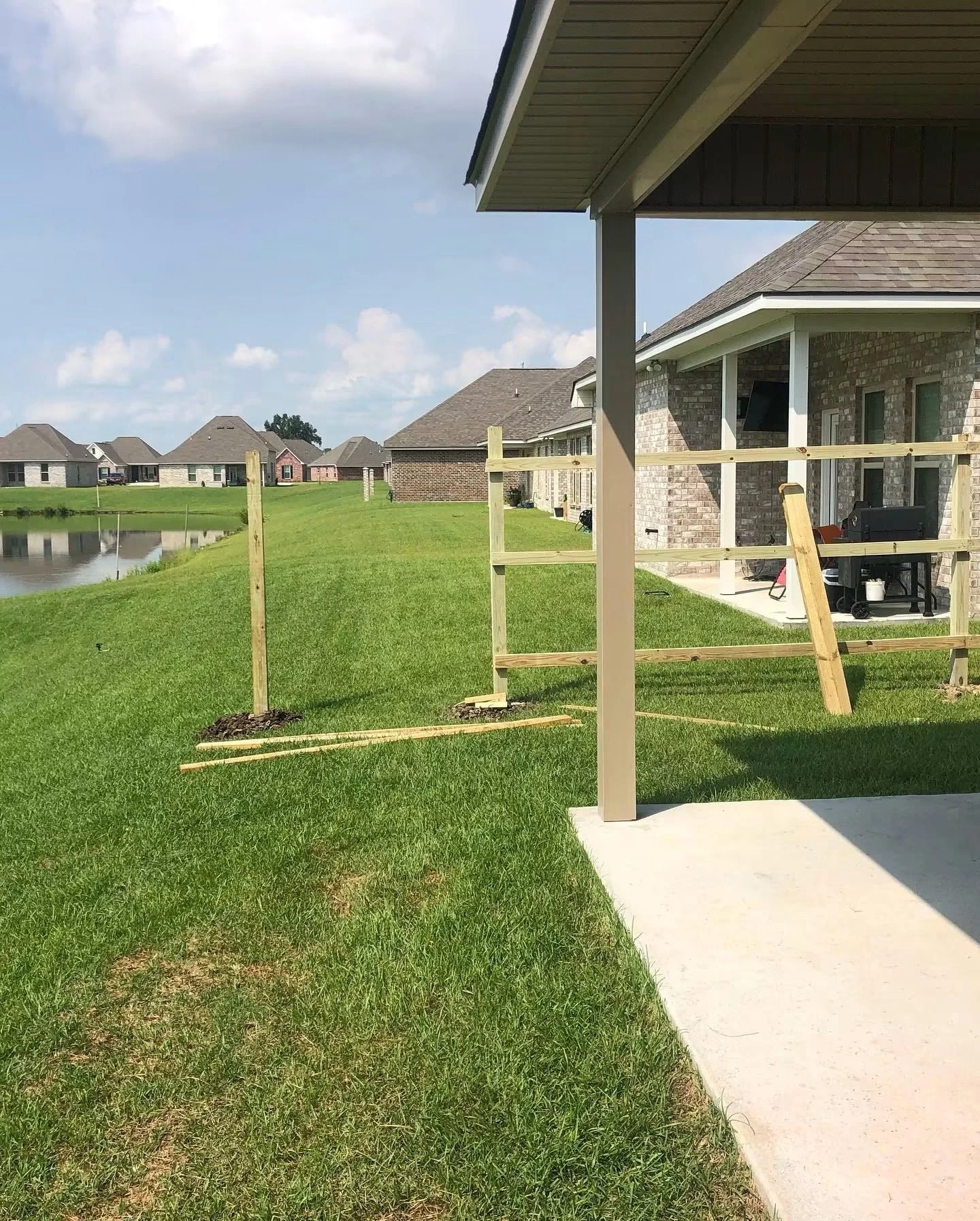 Backyard with a new wooden railing, grass, and a concrete patio. Several houses are visible across a small lake or pond, under a partly cloudy sky.