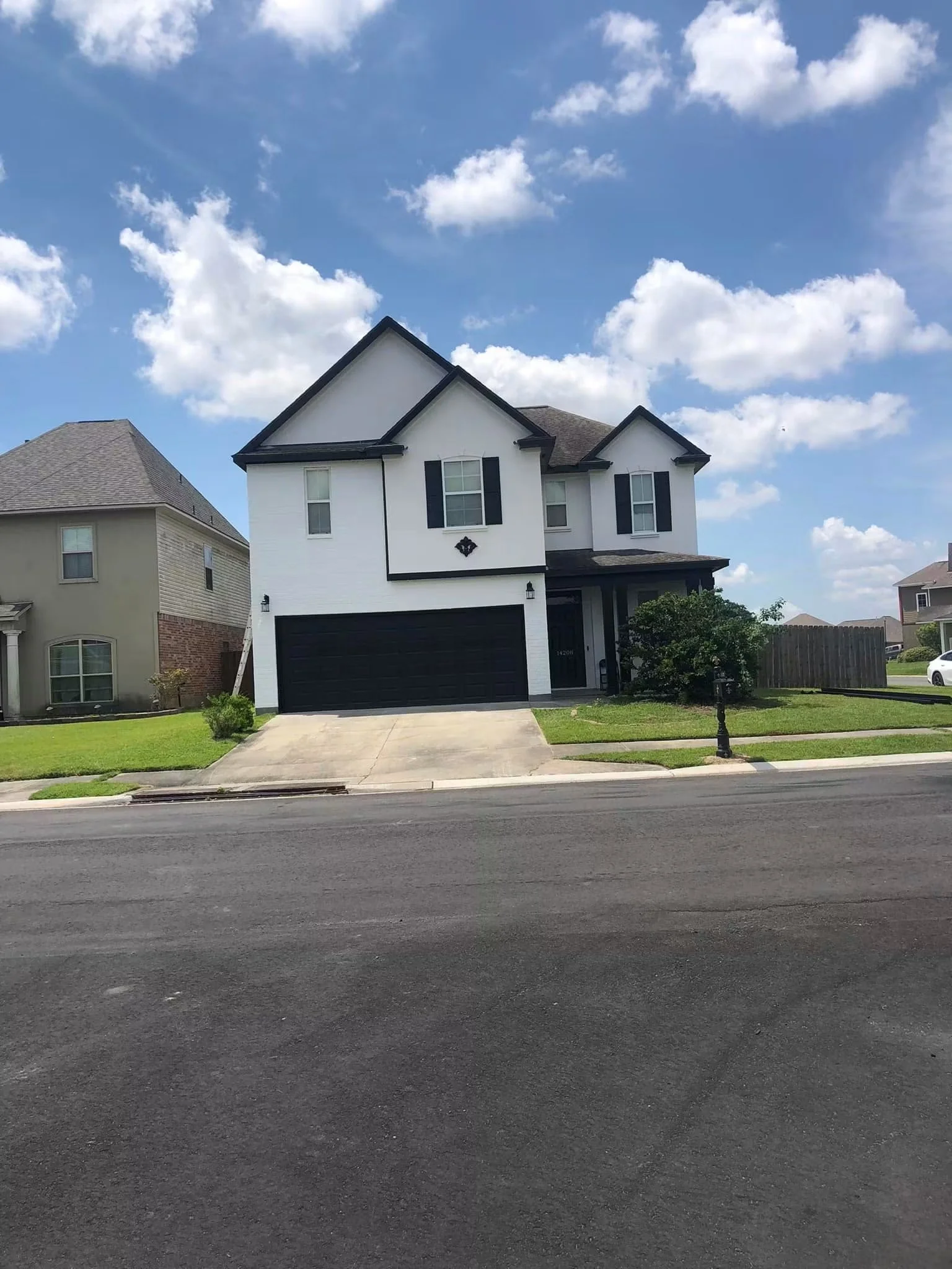 Front view of a two-story white house with black shutters, a black garage door, a driveway, a small lawn, and a wooden fence. The sky is partly cloudy with white clouds and blue sky.