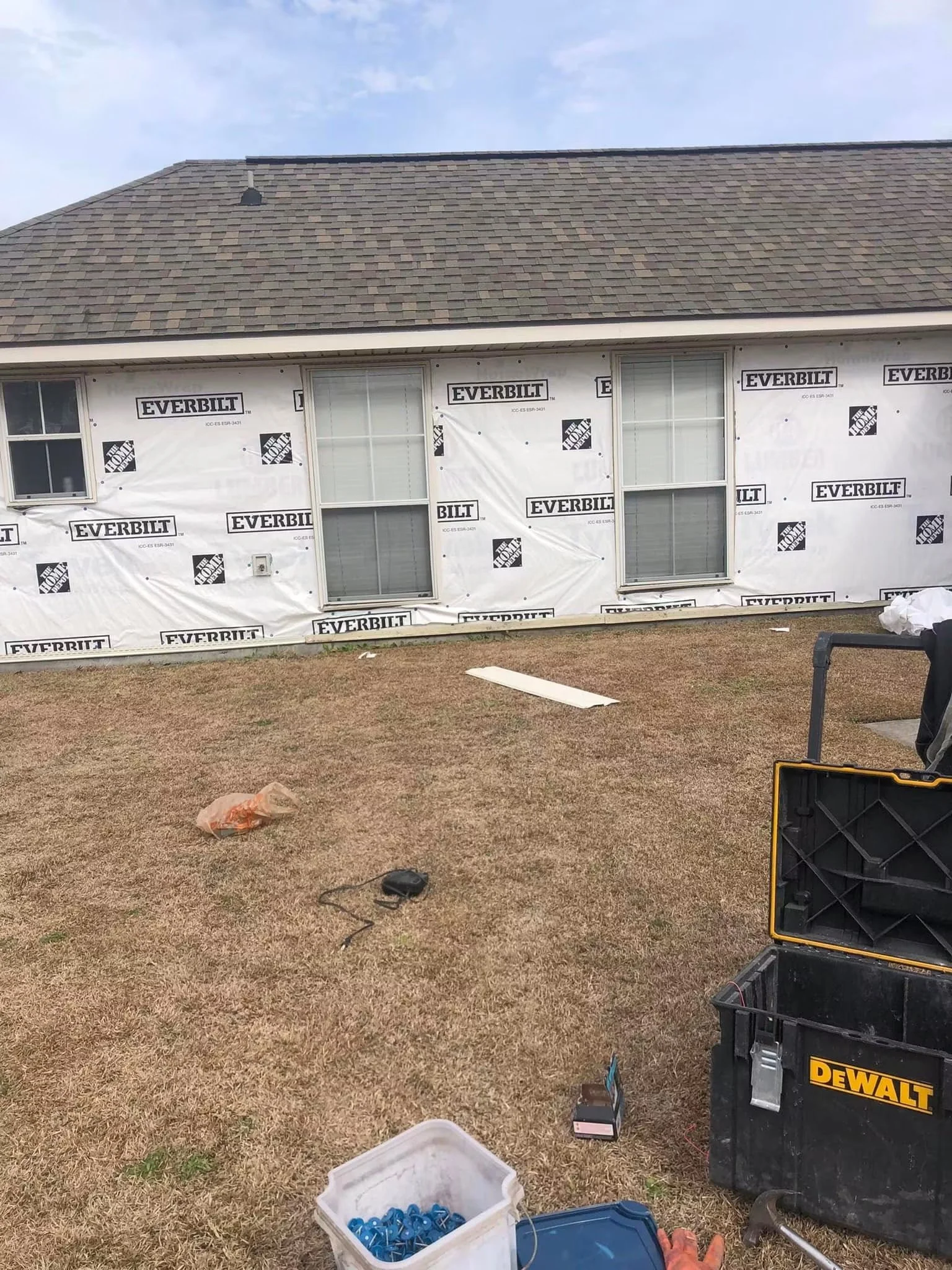 Construction site with a house under renovation, covered in GeoBilt house wrap, and tools and supplies scattered on dry grass in the foreground.