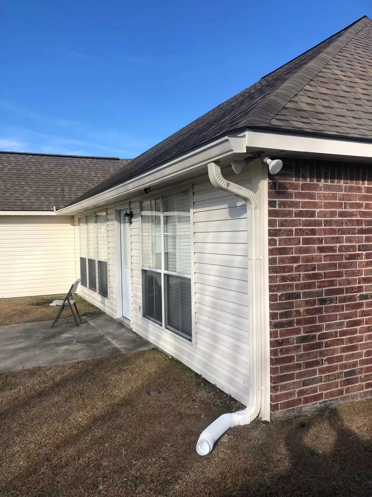 The image shows the corner of a house with a roof, brick wall, and a sliding glass door. There is a white gutter with a downspout and a white flexible drain pipe extending from the gutter to the ground. A small black lamp is mounted on the wall next 