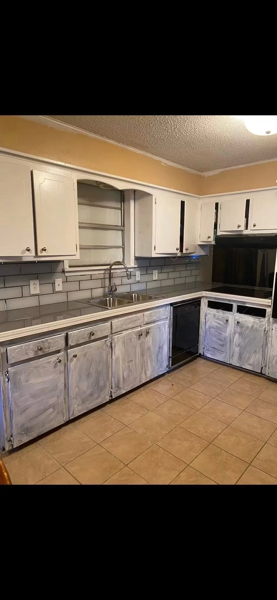 Kitchen with white upper and distressed lower cabinets, gray tile backsplash, double sink, black appliances, and tiled floor.