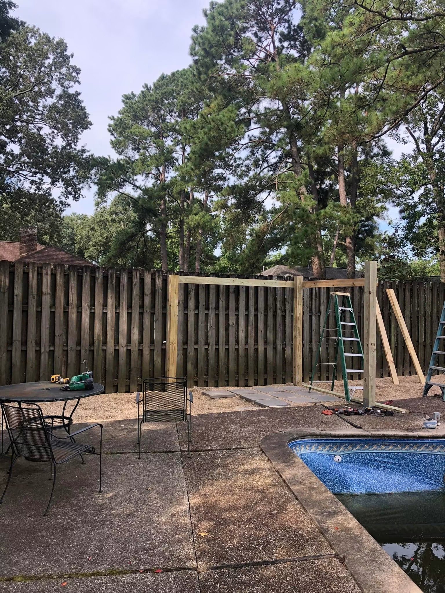 Backyard with a wooden fence, partially built structure, outdoor furniture, a small swimming pool, and tall trees in the background.