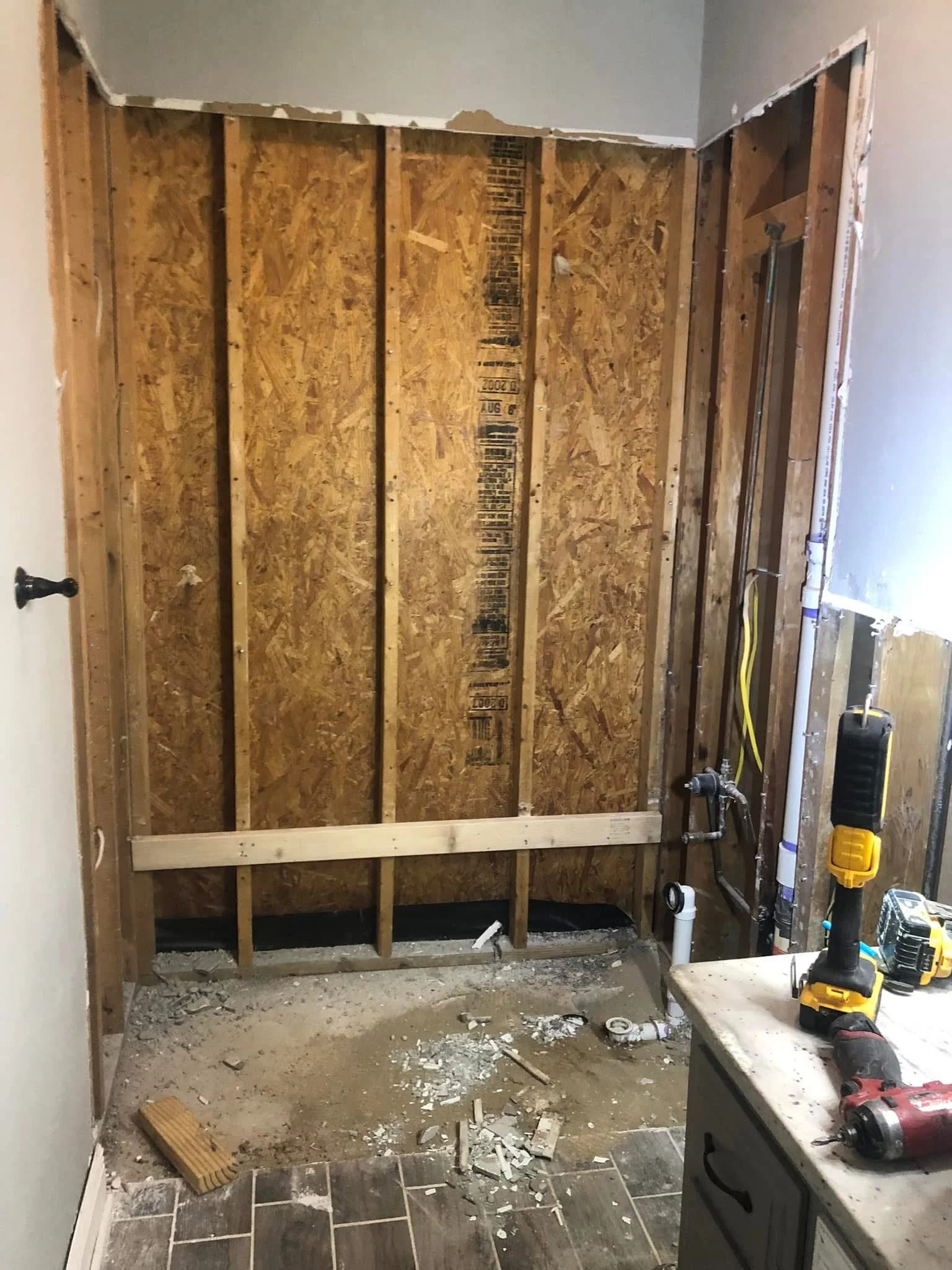 Bathroom in the process of renovation with a partially removed wall, showing exposed wooden studs and plumbing pipes, a workbench with tools, and debris on the floor.