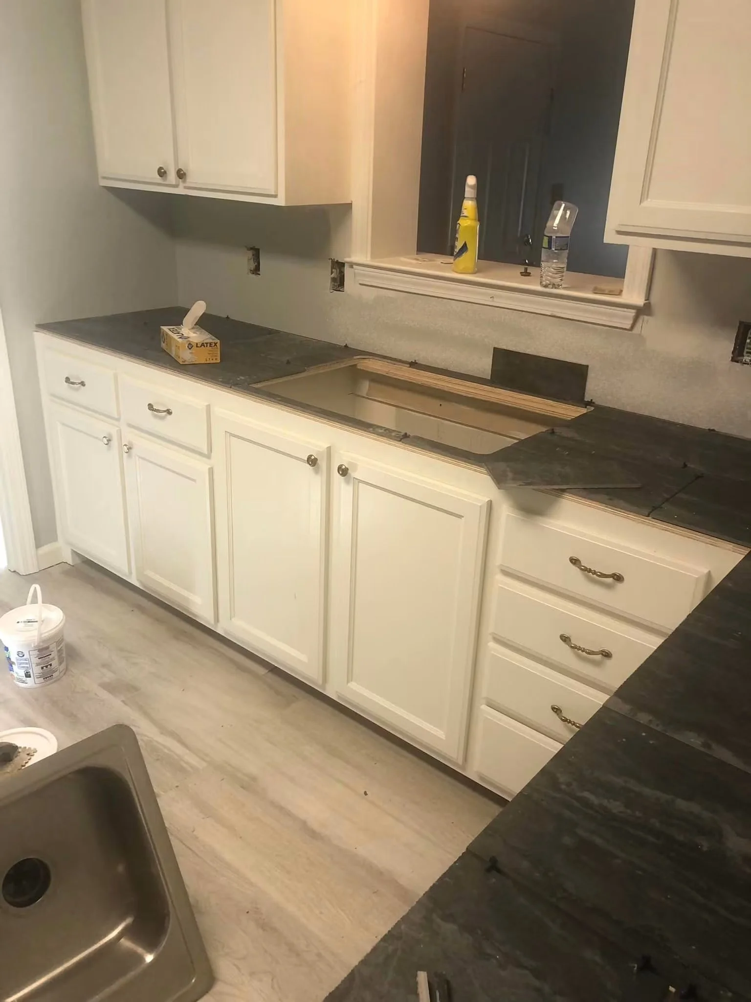 Kitchen under renovation with white cabinets, black countertops, and construction supplies visible, including a box of latex gloves and a paint bucket.