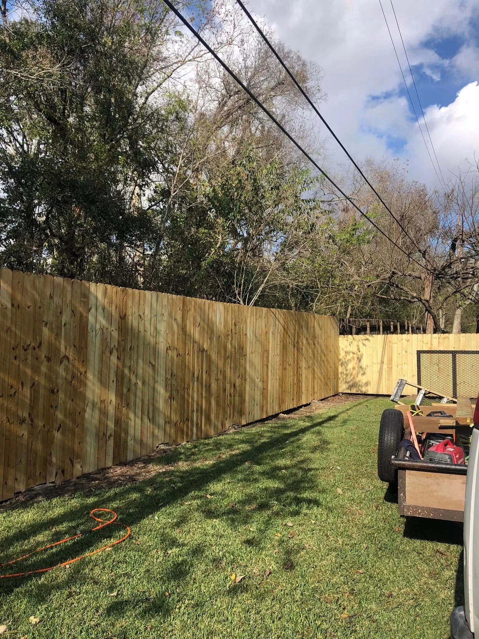 A backyard shows a newly installed wooden fence, with some backyard tools and equipment on a small cart in the foreground, and trees with some leaves and some without, under partly cloudy sky.