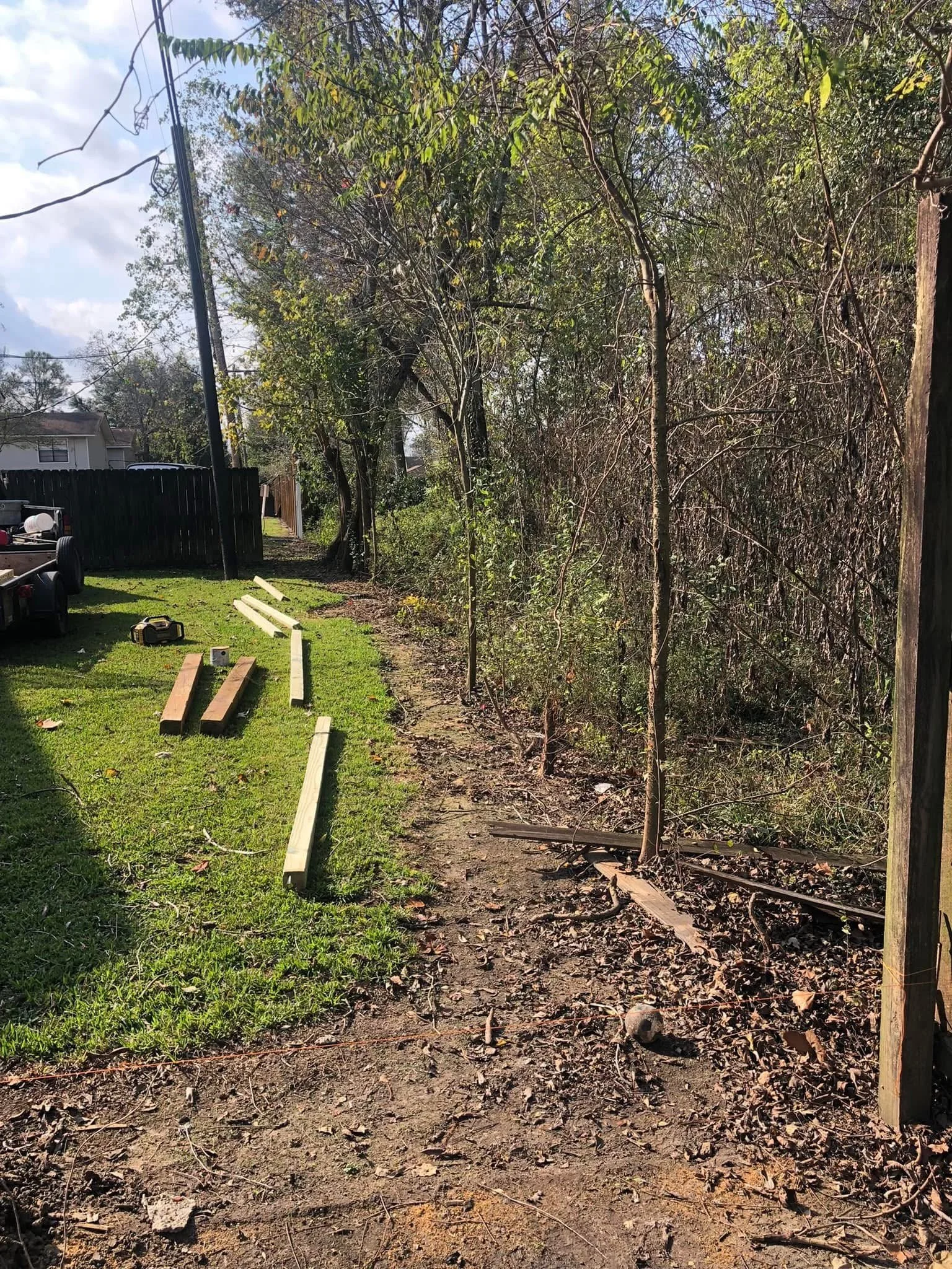 A backyard with a grassy area and a pathway under construction with wooden planks and tools, partially enclosed by a wooden fence and lined with trees and bushes.