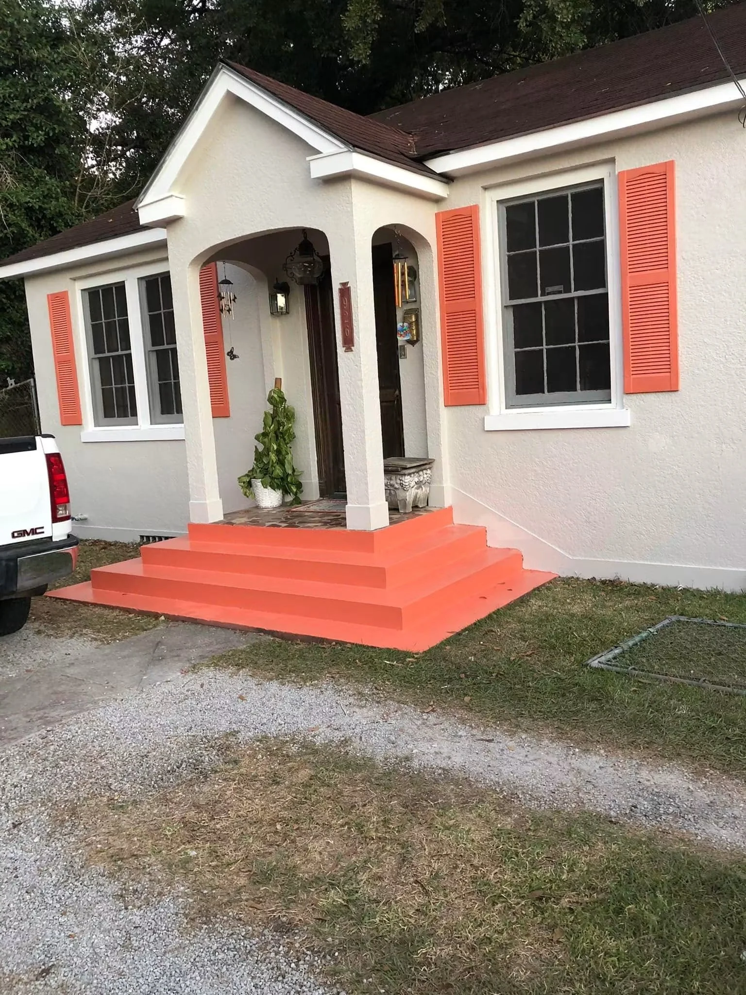 Front entrance of a white house with orange shutters, concrete steps painted orange, and a small porch with decorations and plants. A white vehicle is partially visible on the left side of the image, and the ground is a mixture of gravel and grass.