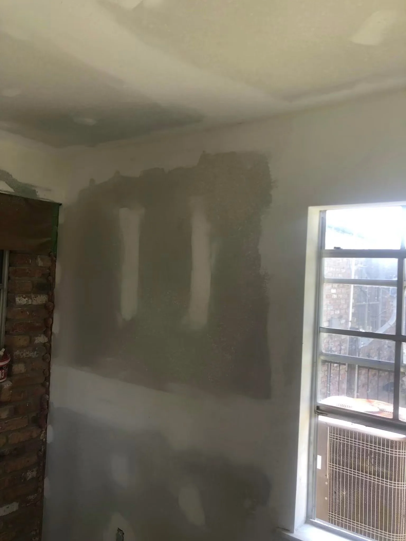Interior of a room under renovation with partially finished walls, exposed brick to the left, and a window on the right side with sunlight coming through.