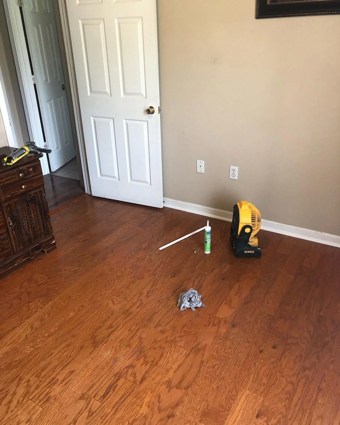 A room with hardwood floor, a slightly open white door leading to a darker room, a wooden dresser on the left, and tools including a yellow work light, a glue bottle, and a rag on the floor.
