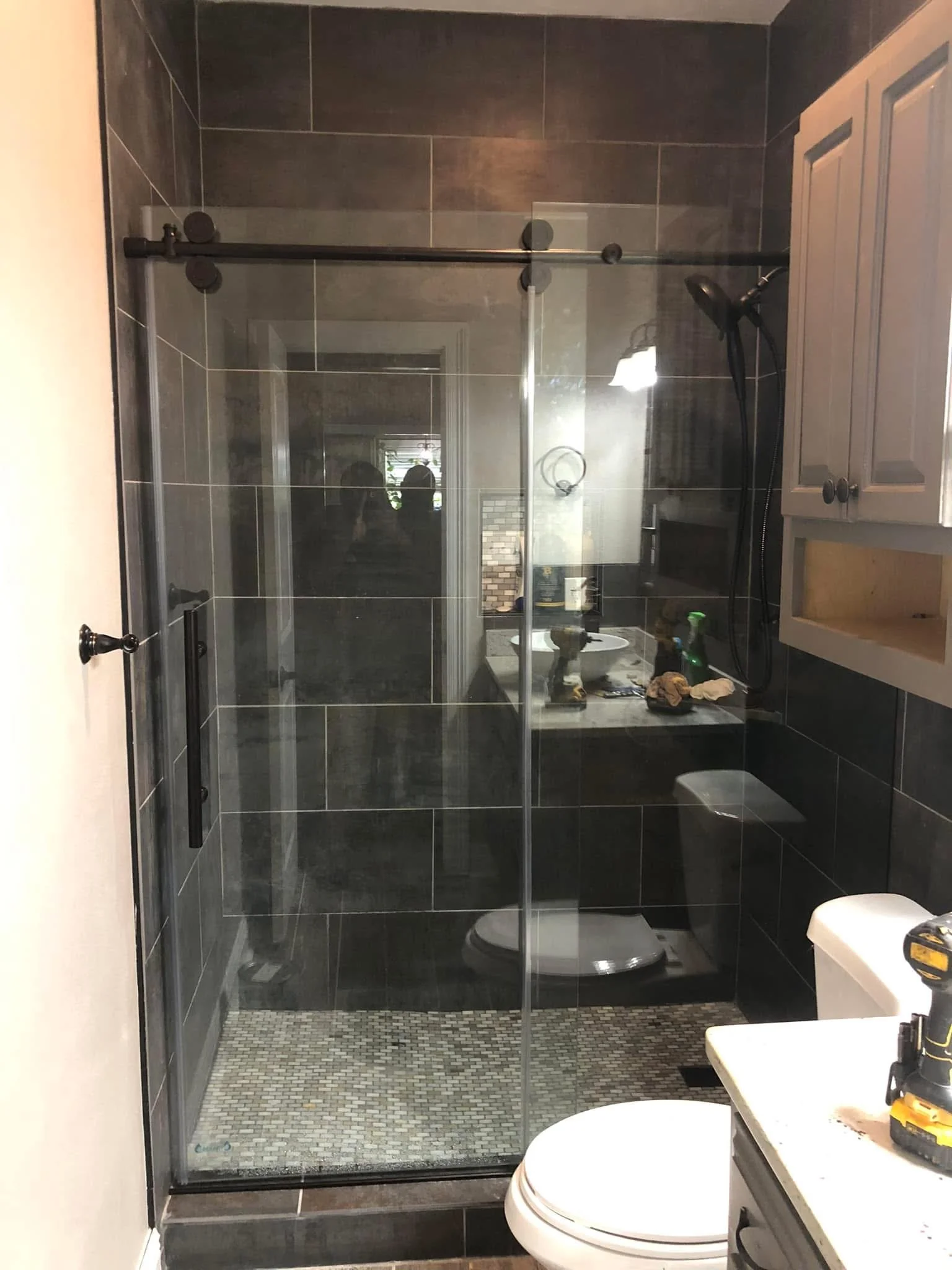 A bathroom with a shower enclosed by glass walls, a dark tile shower area, a small mosaic tile floor, and a toilet. The reflection in the glass shows part of the room and some objects on the countertop.