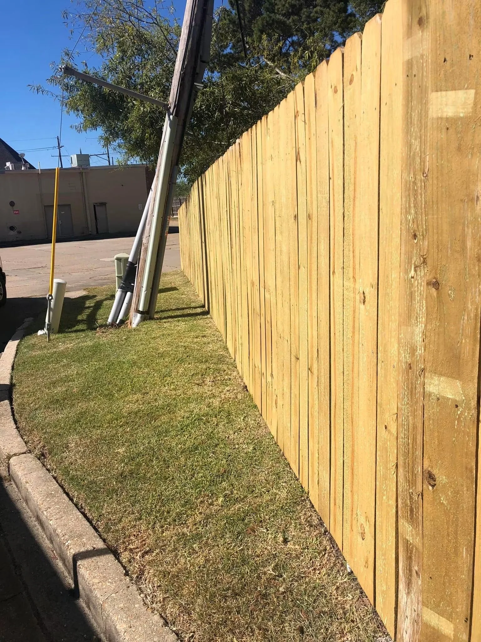 A newly installed wooden fence along a sidewalk with green grass and a curb, in an outdoor area under a clear blue sky.