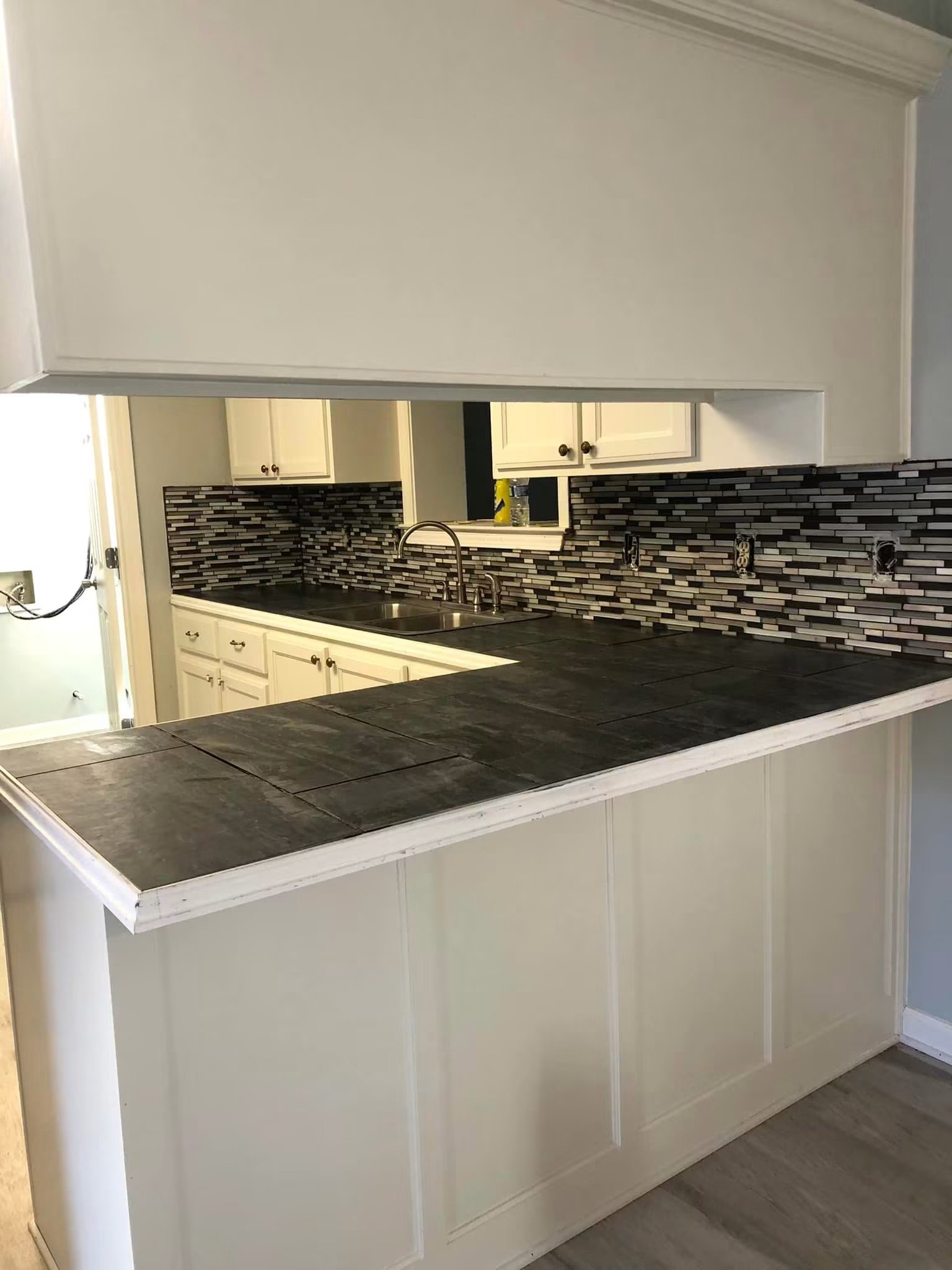Kitchen with white cabinets, black countertops, mosaic tile backsplash, and a stainless steel sink, seen from an island or breakfast bar.