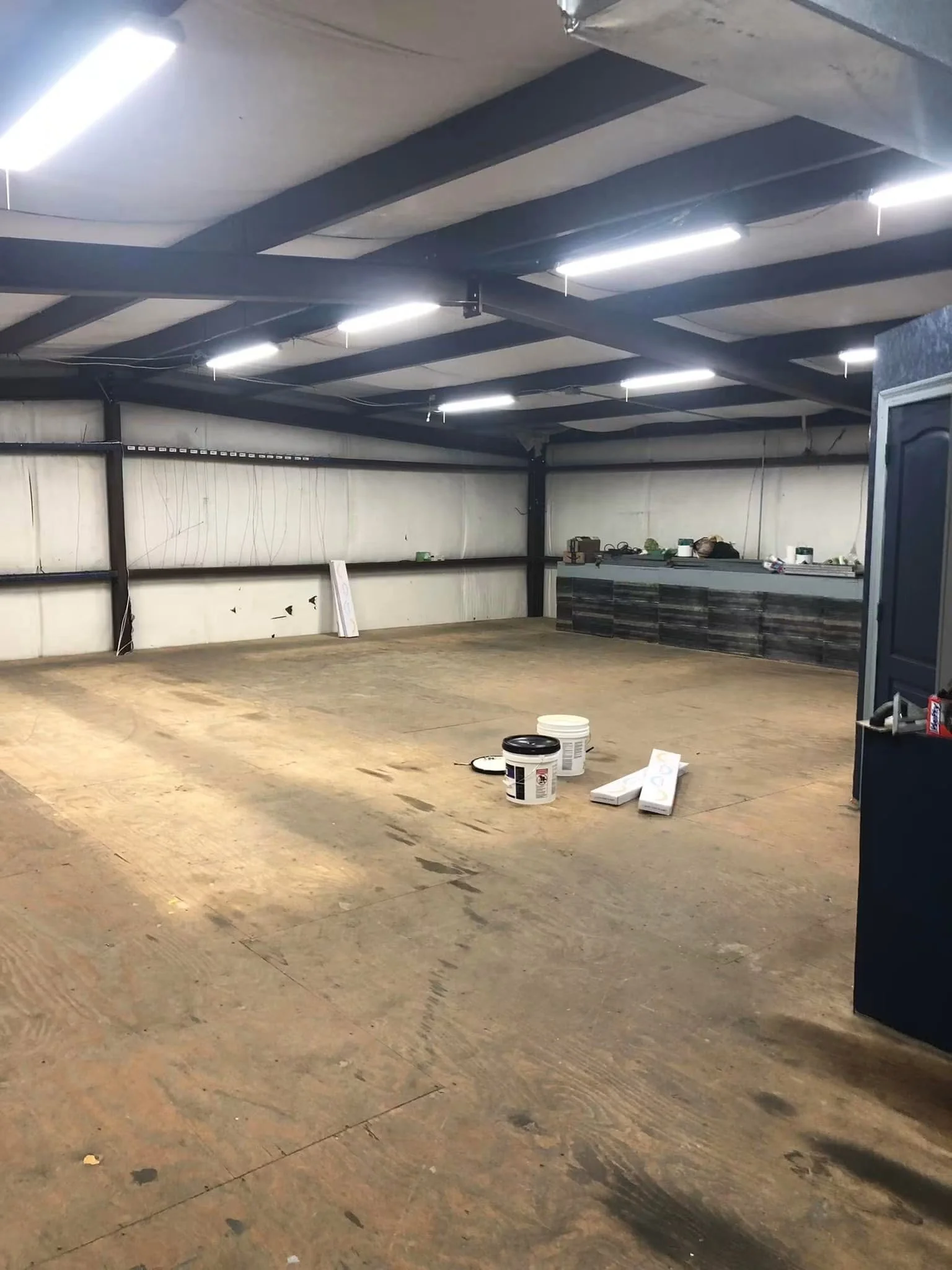 Empty indoor space with a concrete floor, metal framework, fluorescent lighting, and some construction materials like paint buckets and drywall.