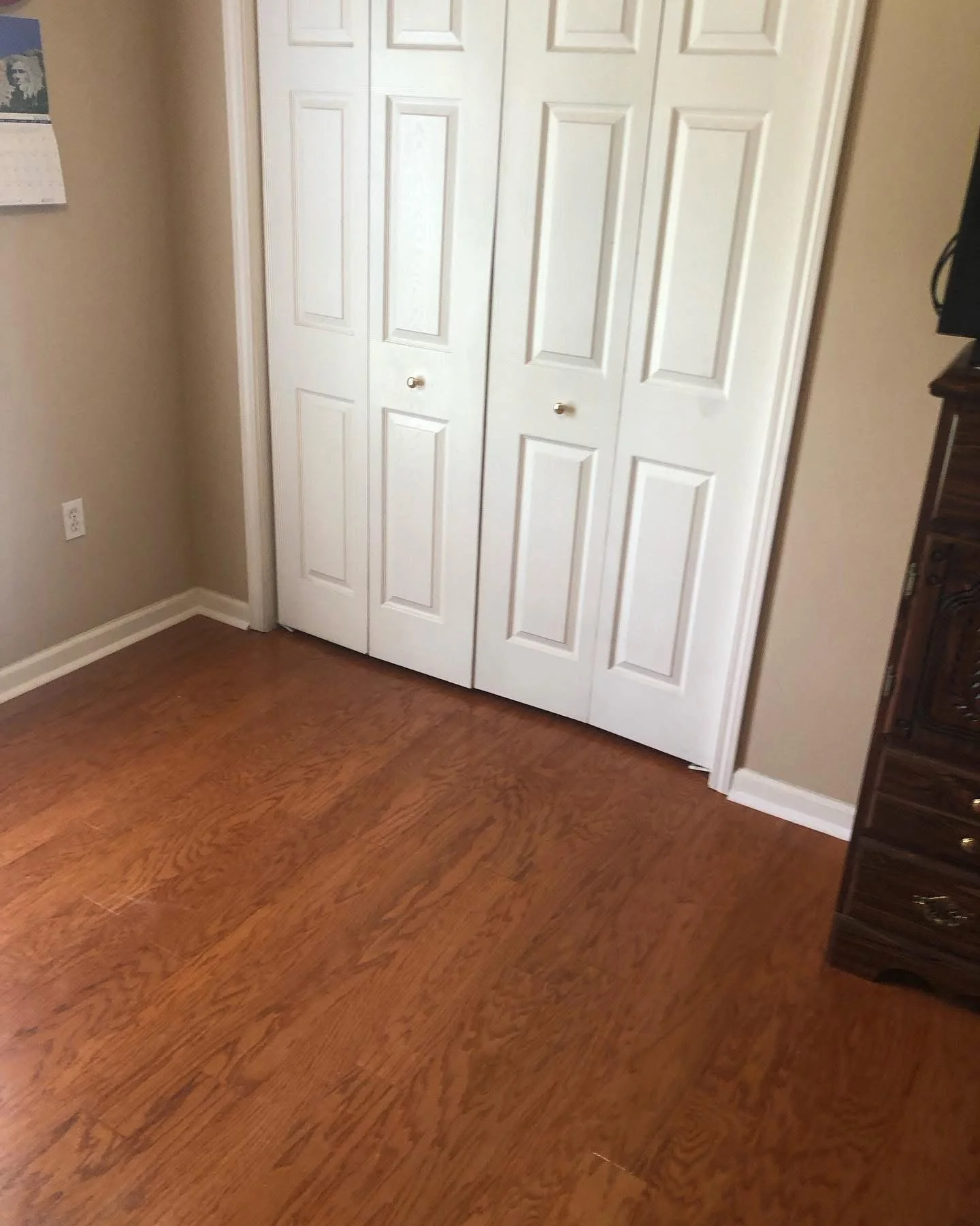 Room with hardwood floors, white closet doors, beige walls, a dark wooden dresser on the right, and a calendar with a black and white image on the wall to the left.