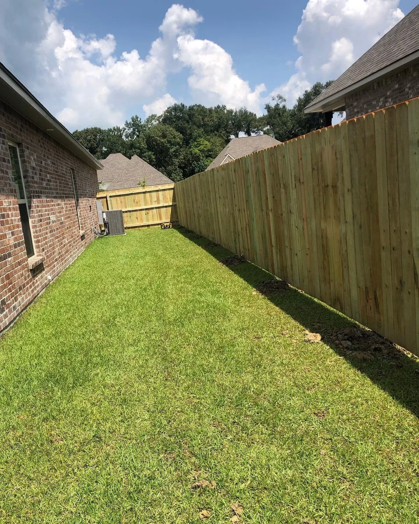 A backyard with freshly laid wood fencing and a well-maintained green lawn. There are neighboring houses with brick walls and shingled roofs. The sky is blue with scattered white clouds.