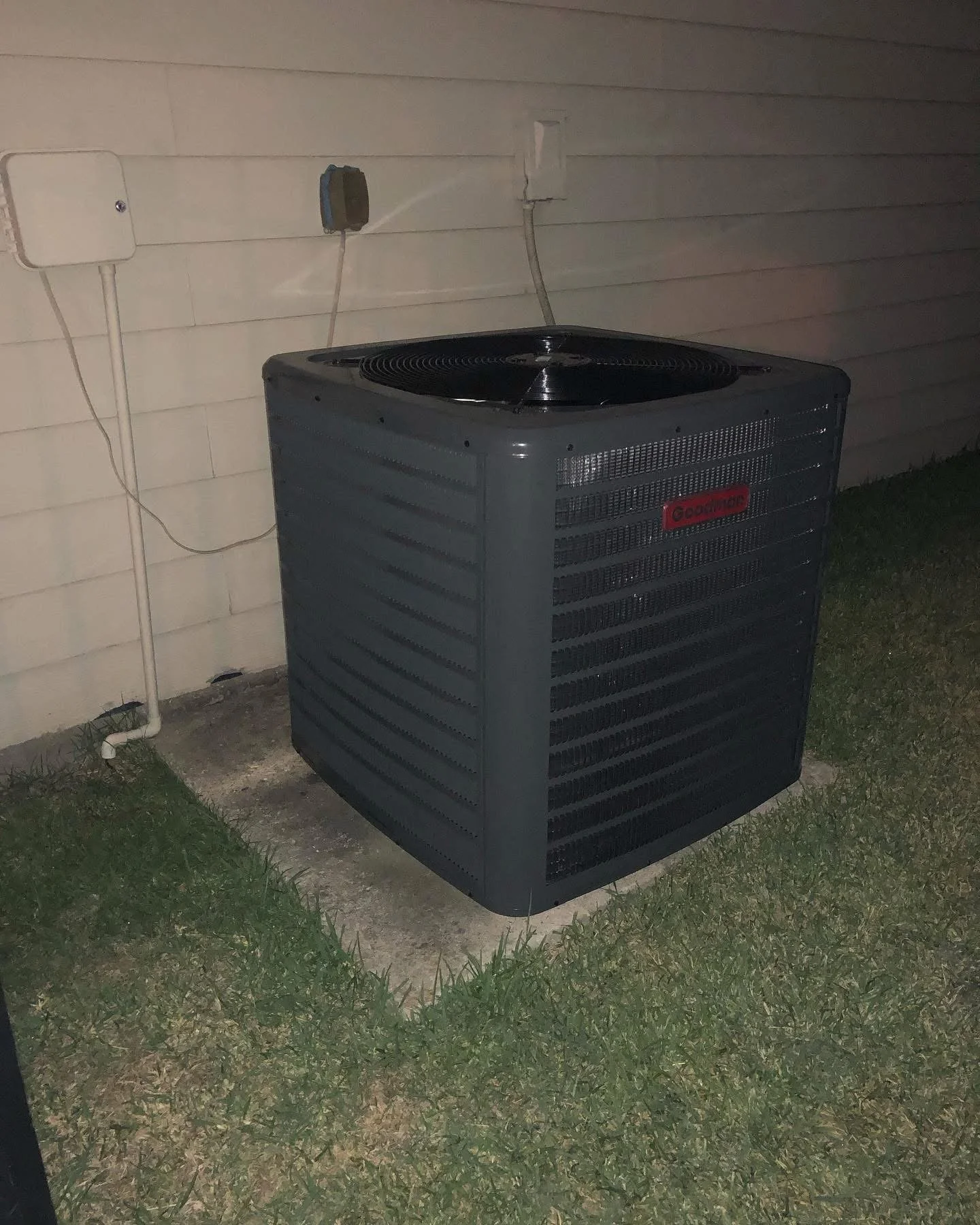 An outdoor air conditioning unit on a concrete pad next to a house wall with electrical wiring and pipes.