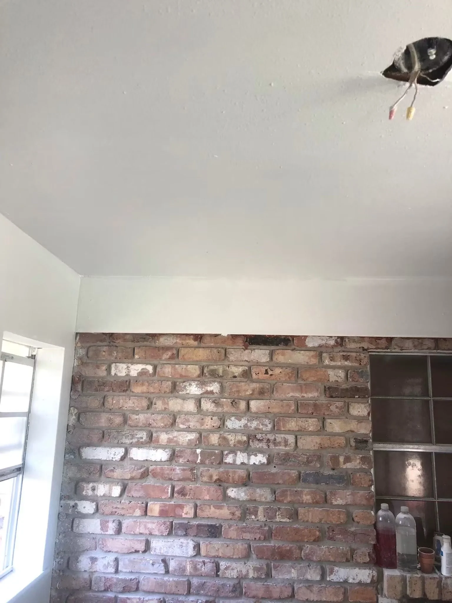 Interior of a room under renovation, showing an exposed brick wall, a white ceiling with a hole for a light fixture, and a window on the left side. There are bottles and cups on a counter in the lower right corner.