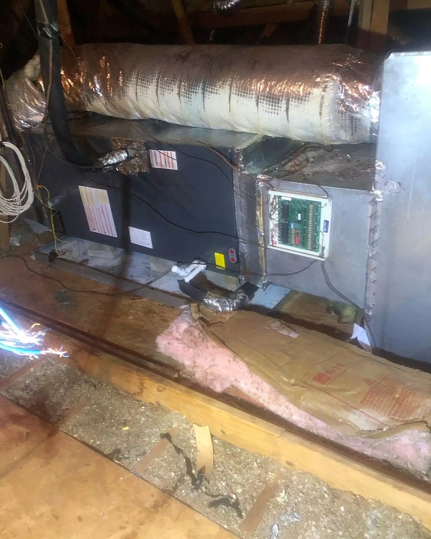 An area of an attic or crawl space showing HVAC ductwork wrapped in insulation and a cooling unit with exposed wiring and components, with insulation and wood framing visible.