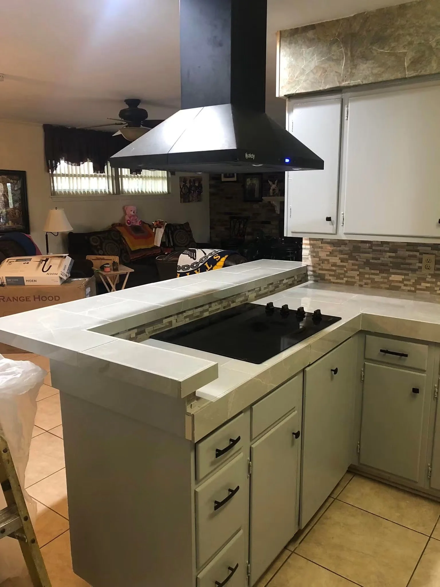 View of a kitchen with white cabinets, beige tiled flooring, a tiled partial wall, a stovetop, and a counter. There is a living room area visible in the background with a black couch, a lamp, and a pink teddy bear on the couch.
