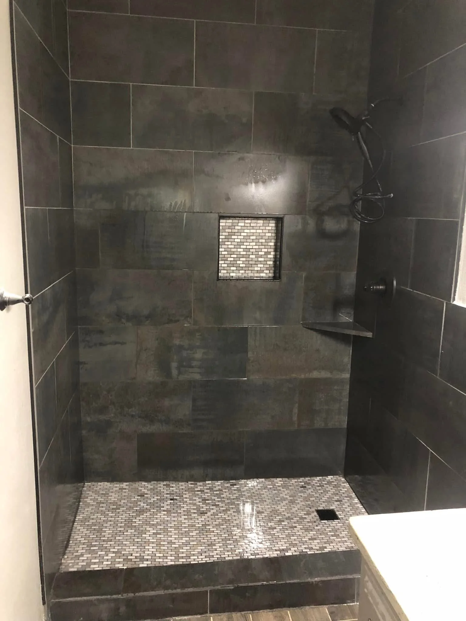 A modern shower area with dark gray large tiles on the walls and small mosaic tiles on the floor, a square drain in the middle, a wall-mounted showerhead, and a soap or shampoo holder.