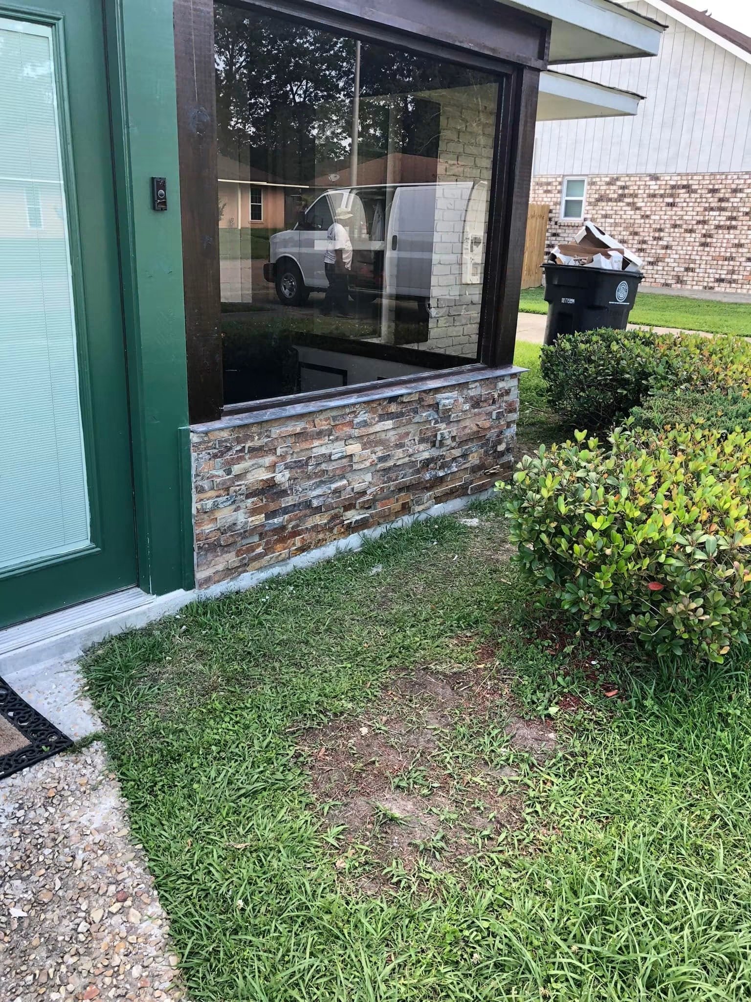 A small front lawn with green grass and a bush next to a brick building with large windows. A gravel path runs alongside the grass. A mailbox and a trash bin are outside the building, reflected in a large window.
