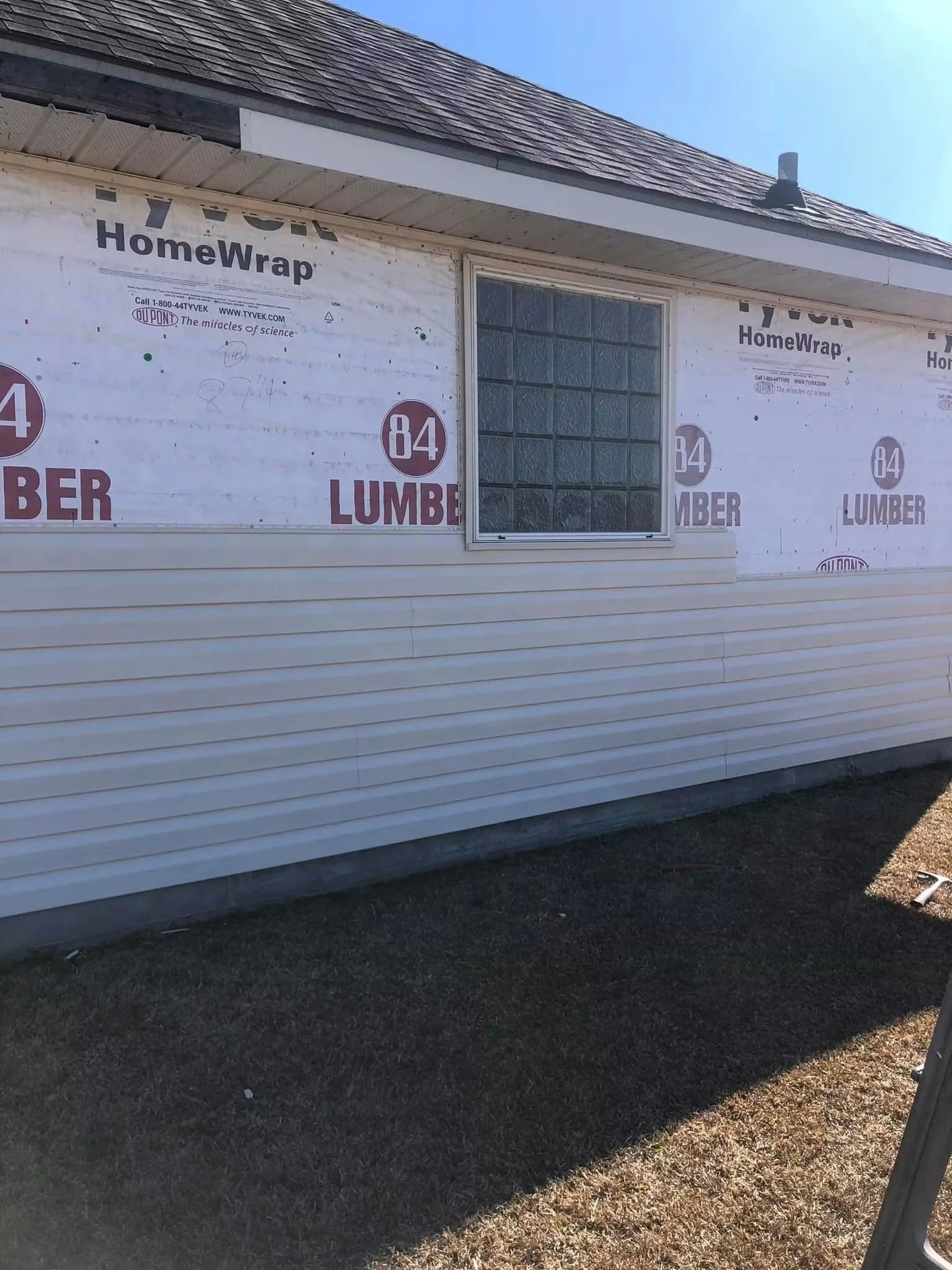 Exterior of a house under construction with HomeWrap insulation material on the walls, a window, and a shingled roof. The house is sided with horizontal white vinyl siding, and there is a shadow cast on the ground.