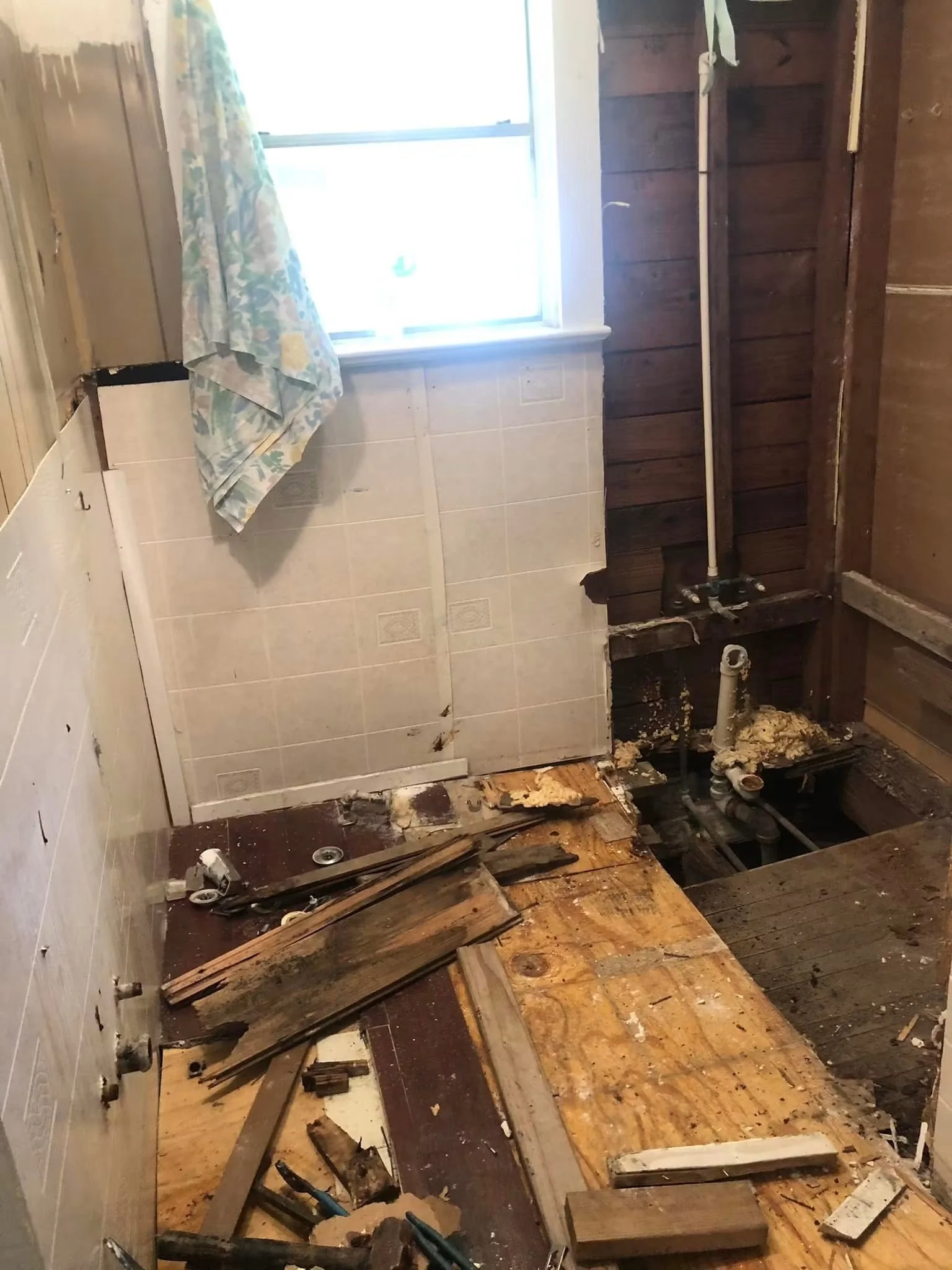 A partially demolished bathroom with exposed pipes, disassembled fixtures, and debris on the floor, with a window and a curtain on the wall.