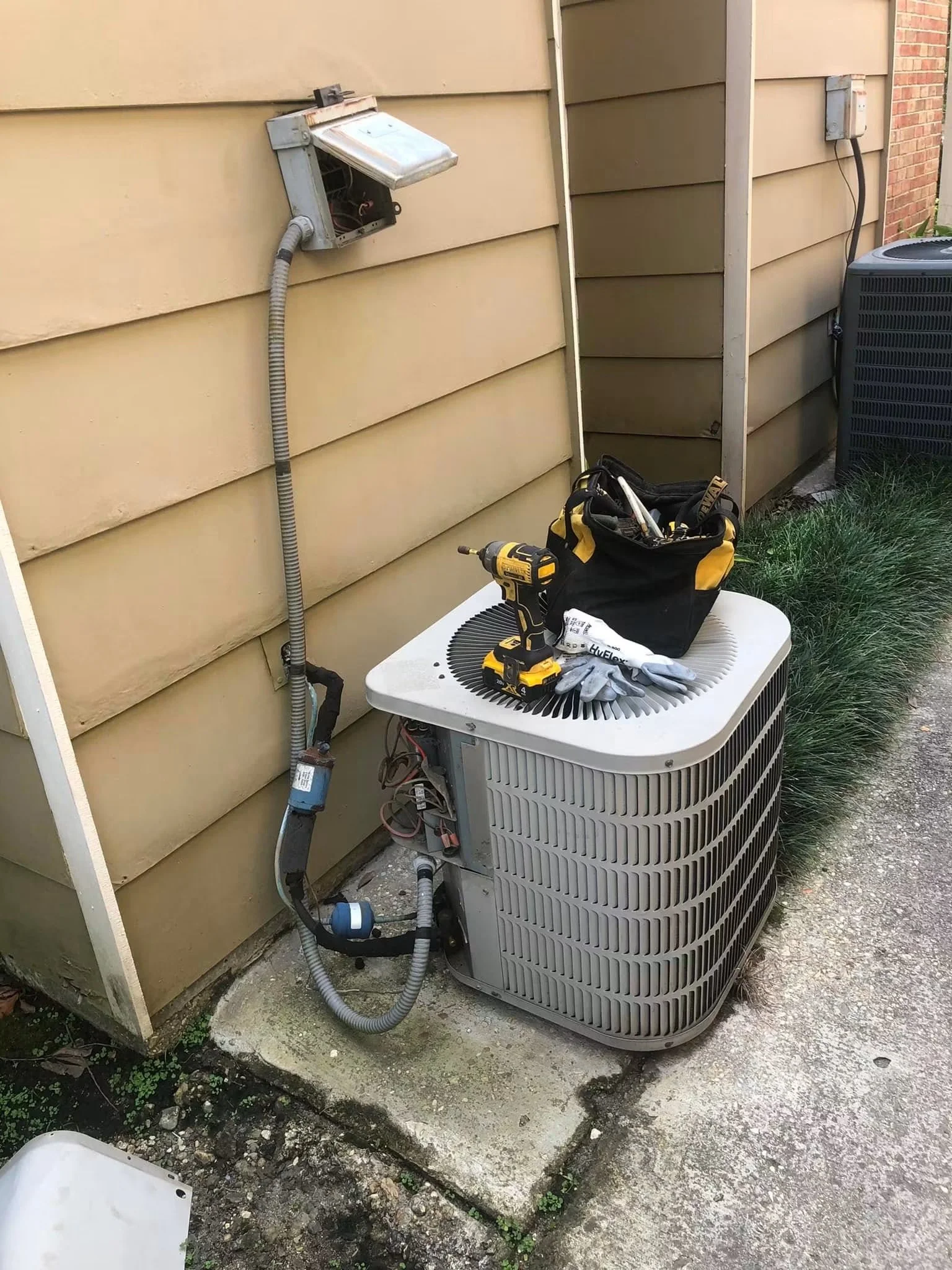 An outdoor HVAC unit with tools and a bag on top, and an opened electrical box with wires, next to a beige house wall and an air conditioning system on the ground.