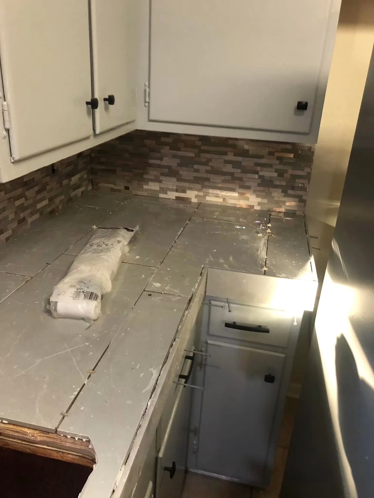 Kitchen with partially installed gray cabinets, a brick-patterned backsplash, and a roll of tile spacers on the floor. The countertop is missing or incomplete.