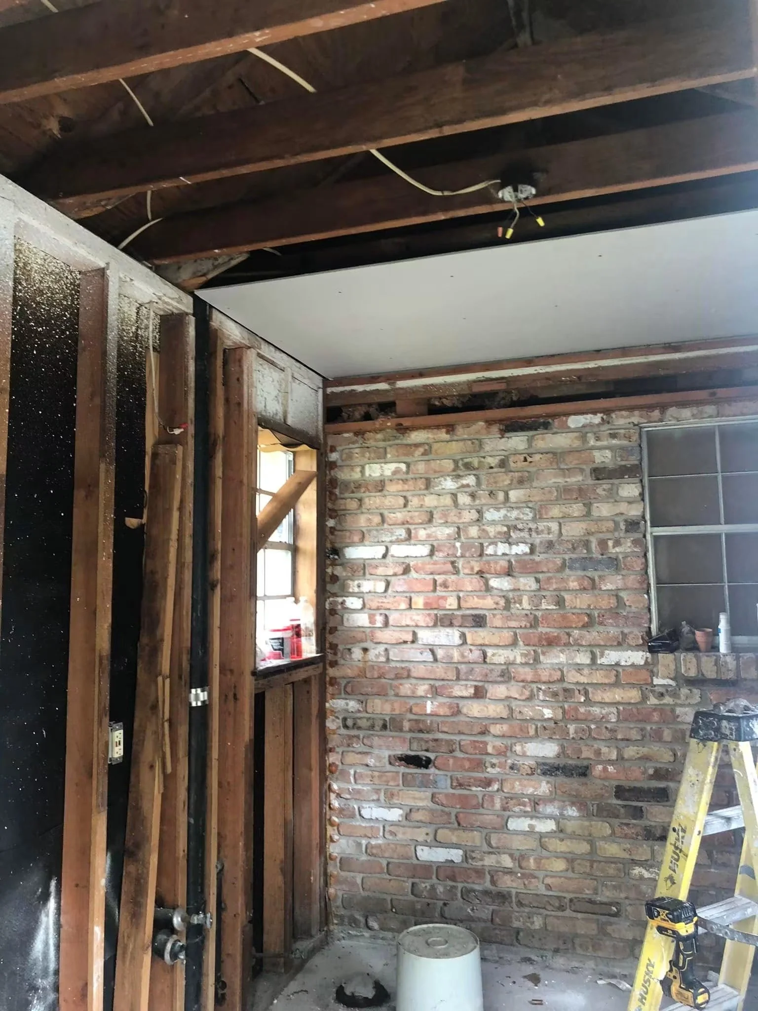 Interior of a room under renovation with exposed brick wall, wooden framing, and a white ceiling panel partially installed. Construction tools and supplies are visible, including a yellow ladder, a drill, and a bucket.