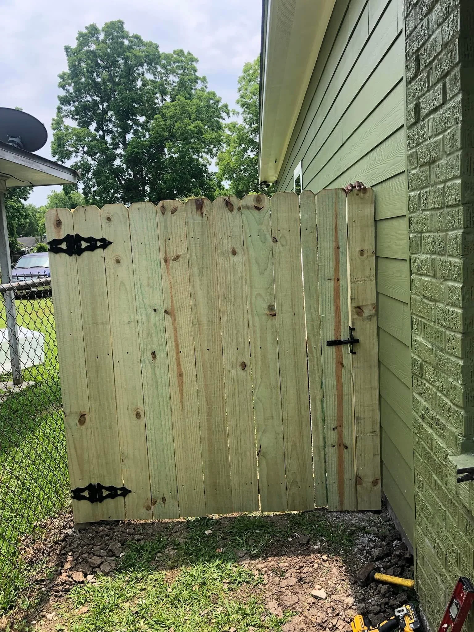 A partially built wooden privacy fence attached to a house with green siding. A door in the fence is held closed with a latch. Some tools, including a cordless drill, are visible on the ground near the fence, and a tree is in the background.