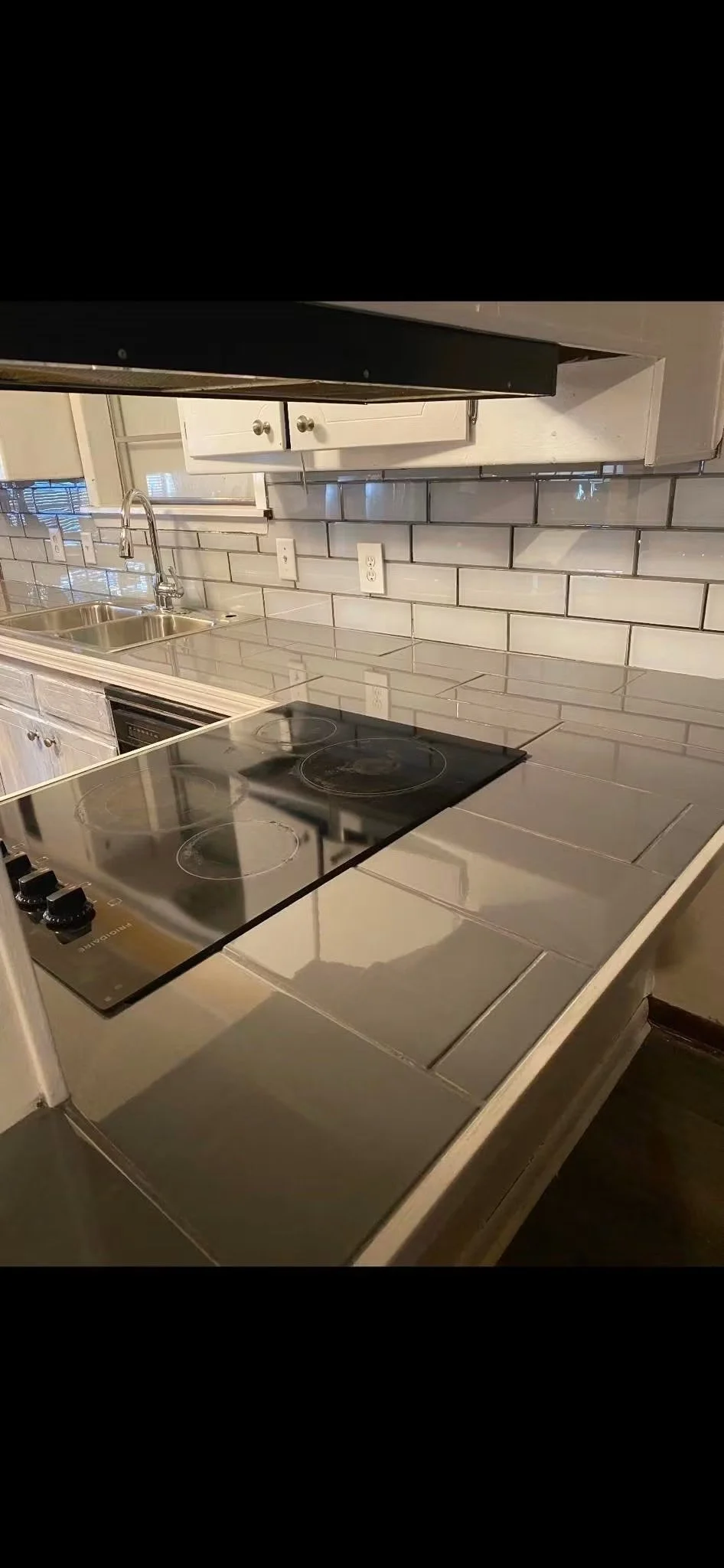 Kitchen with stainless steel countertop, black electric stovetop, white cabinets, gray subway tile backsplash, and a window with blinds above the sink.
