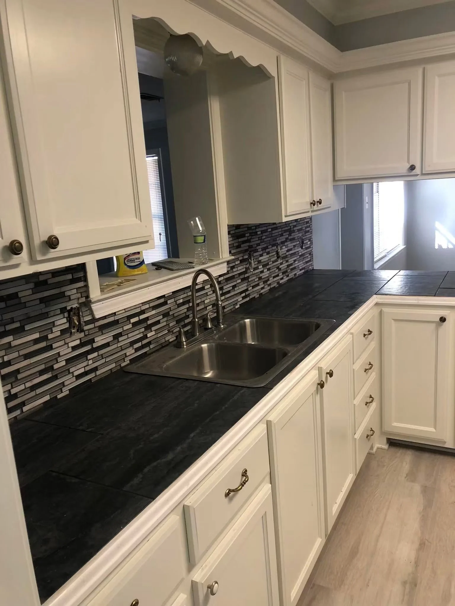 Kitchen with white cabinets, dark countertop, mosaic tile backsplash, double sink, water bottle, scattered papers, and a window with blinds.